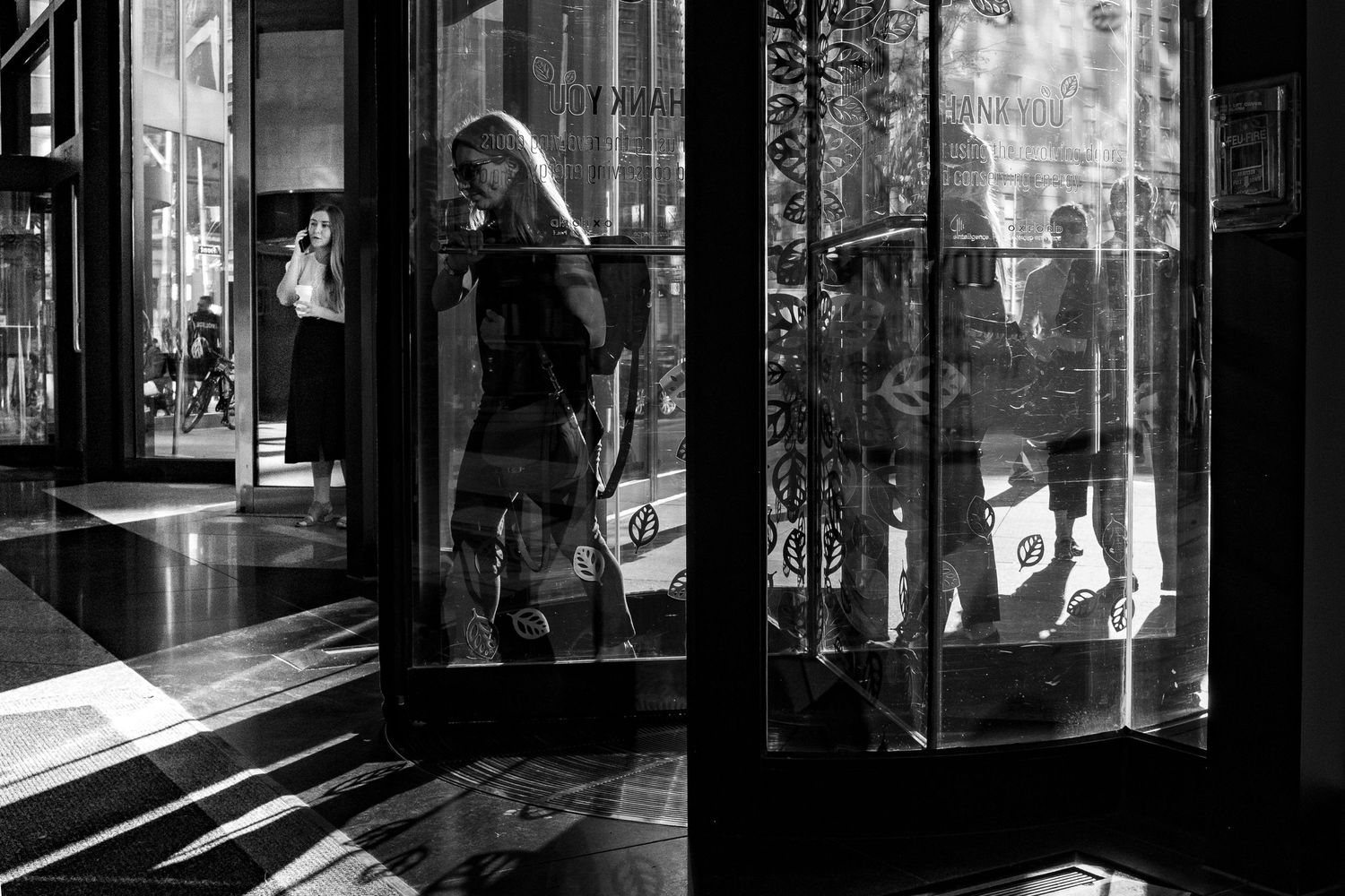 Black and white street photo of a woman walking past a reflective glass storefront door in an urban setting.