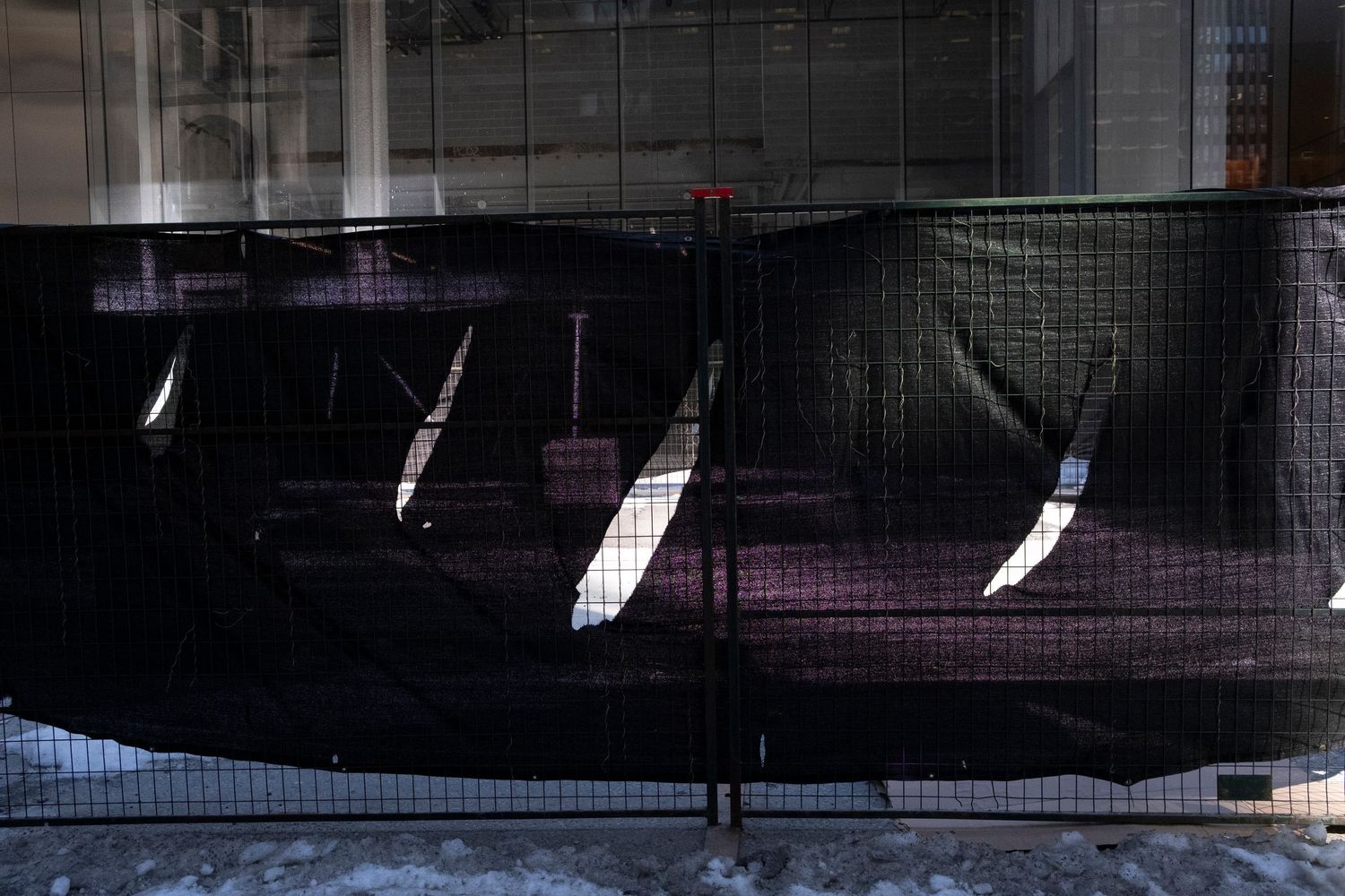 Black mesh construction fence barrier with reflective light patterns on urban sidewalk with snow.