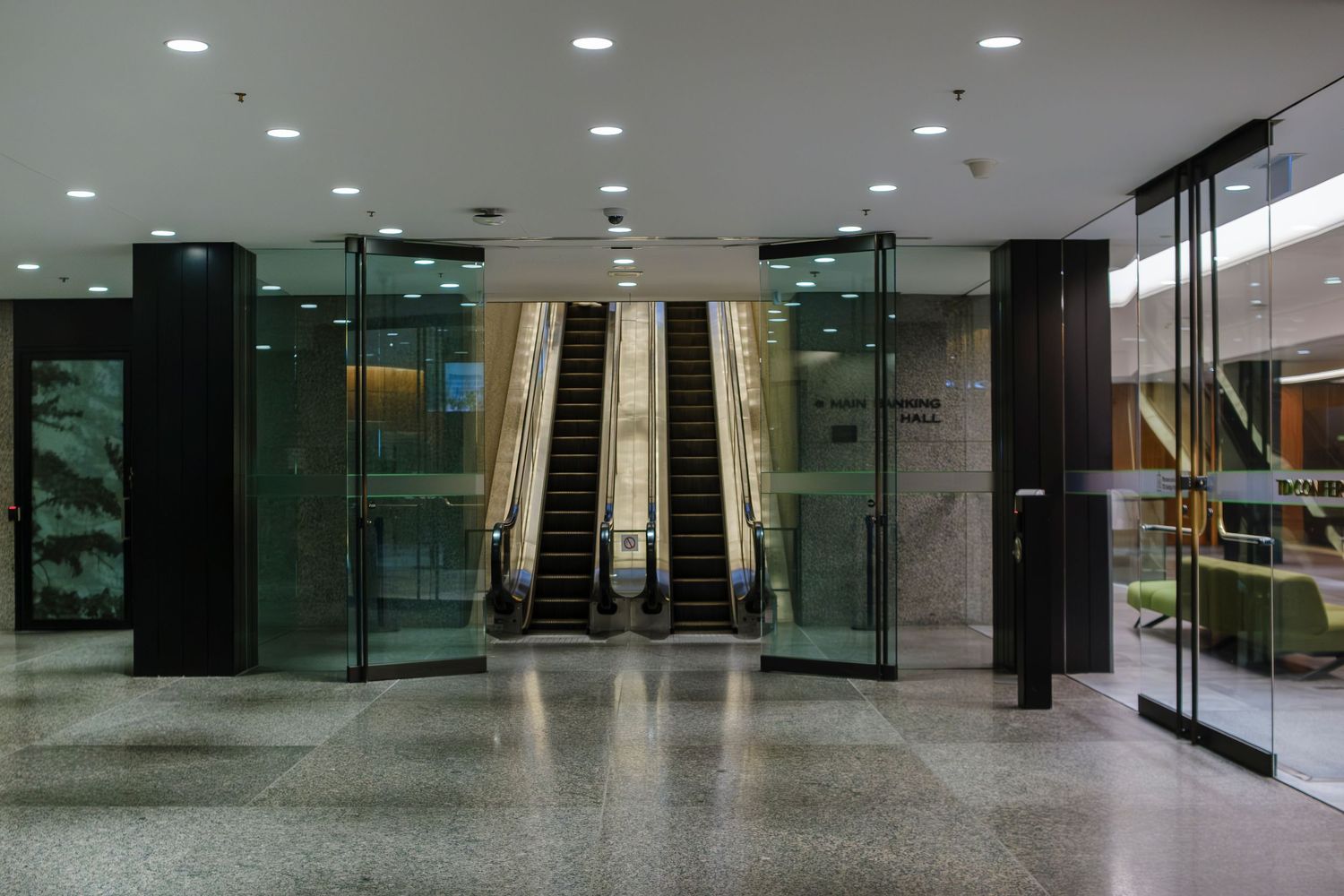 Modern building lobby with escalators, glass doors, marble floors, and recessed ceiling lights in a corporate interior.