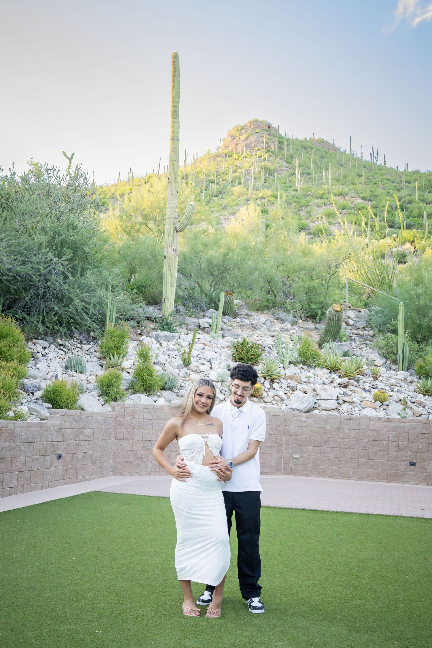 Couple posing together on green lawn with saguaro cactus hillside backdrop during maternity photo session.