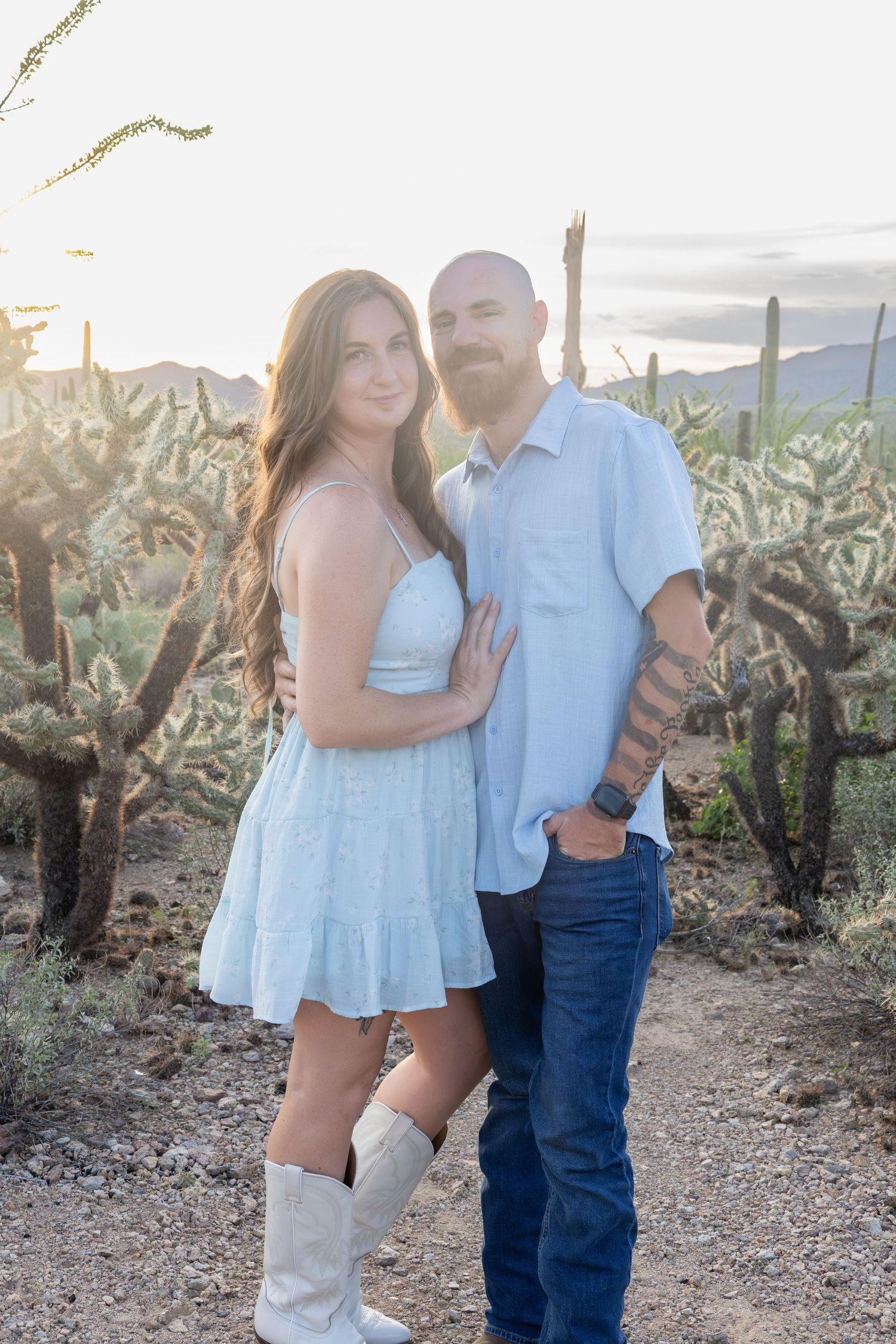 Couple posing together at golden hour in a desert landscape with saguaro cacti and mountains in the background.