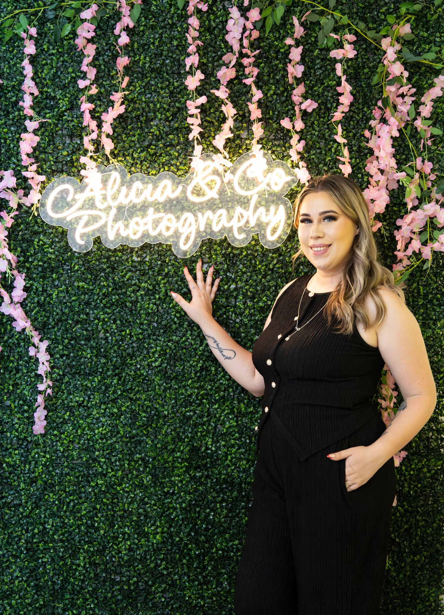 Woman in black jumpsuit posing beside Alicia & Co Photography neon sign on green floral wall backdrop.