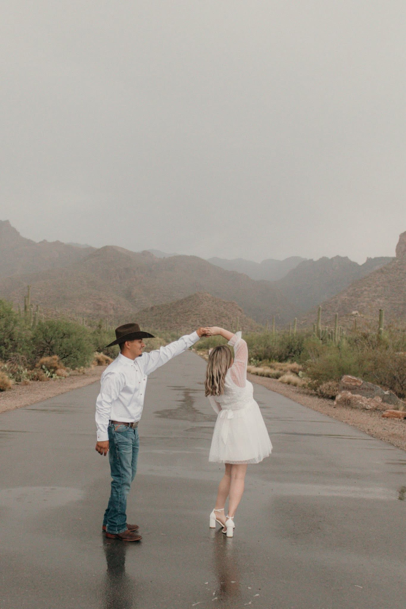 Couple dancing on wet desert road with saguaro cacti and mountains in misty background during rain.