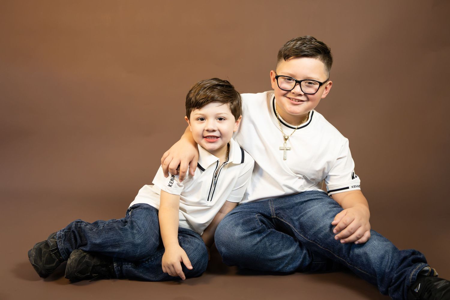 Two young brothers in white shirts and jeans pose together smiling against a warm brown studio background.