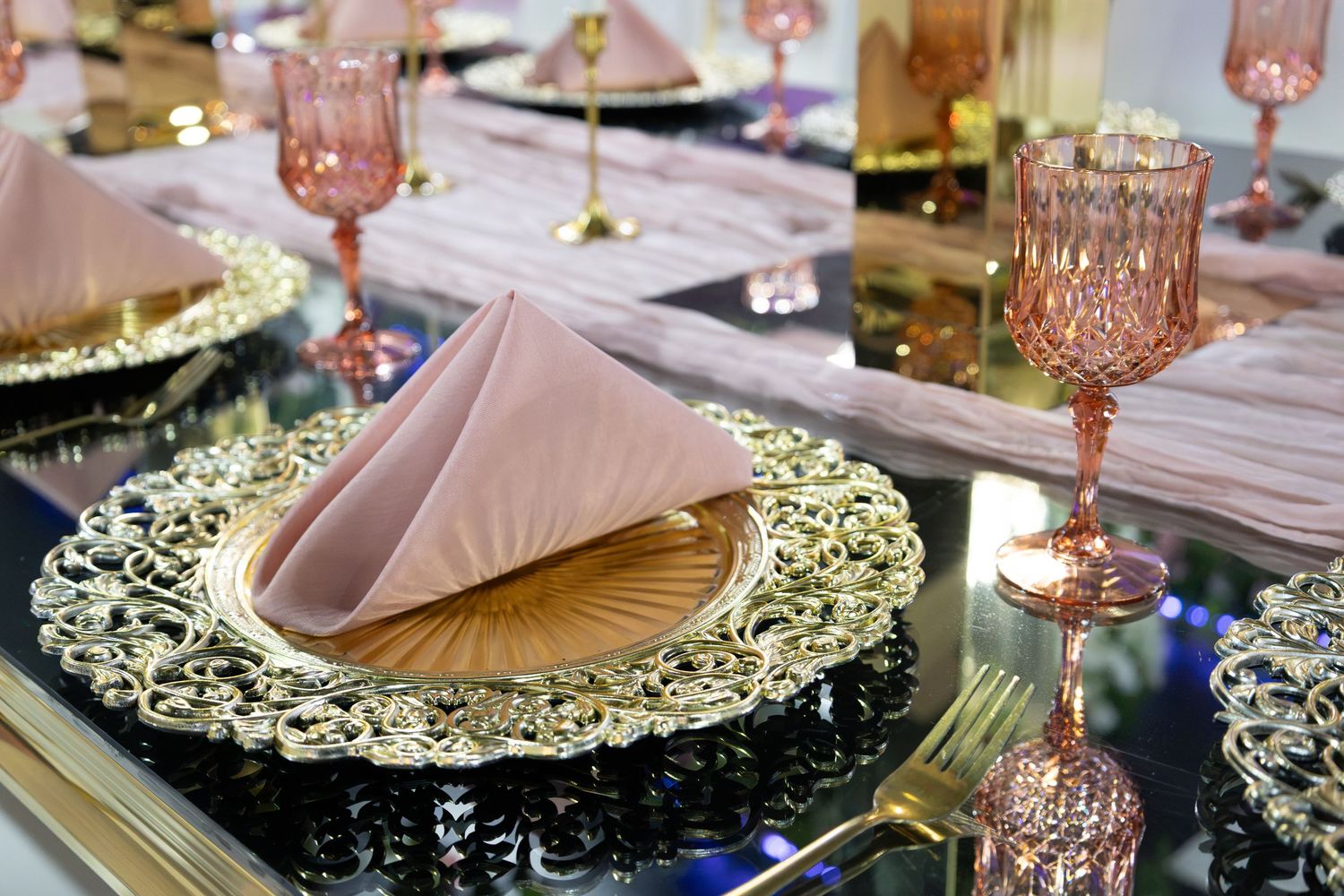 Elegant gold charger plates with pink folded napkins and rose-colored crystal glasses on mirrored table.