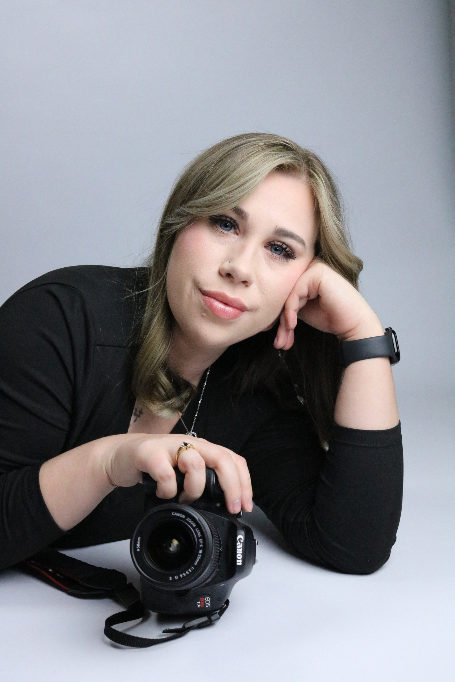 Blonde female photographer posing with a Canon DSLR camera against a gray studio background.
