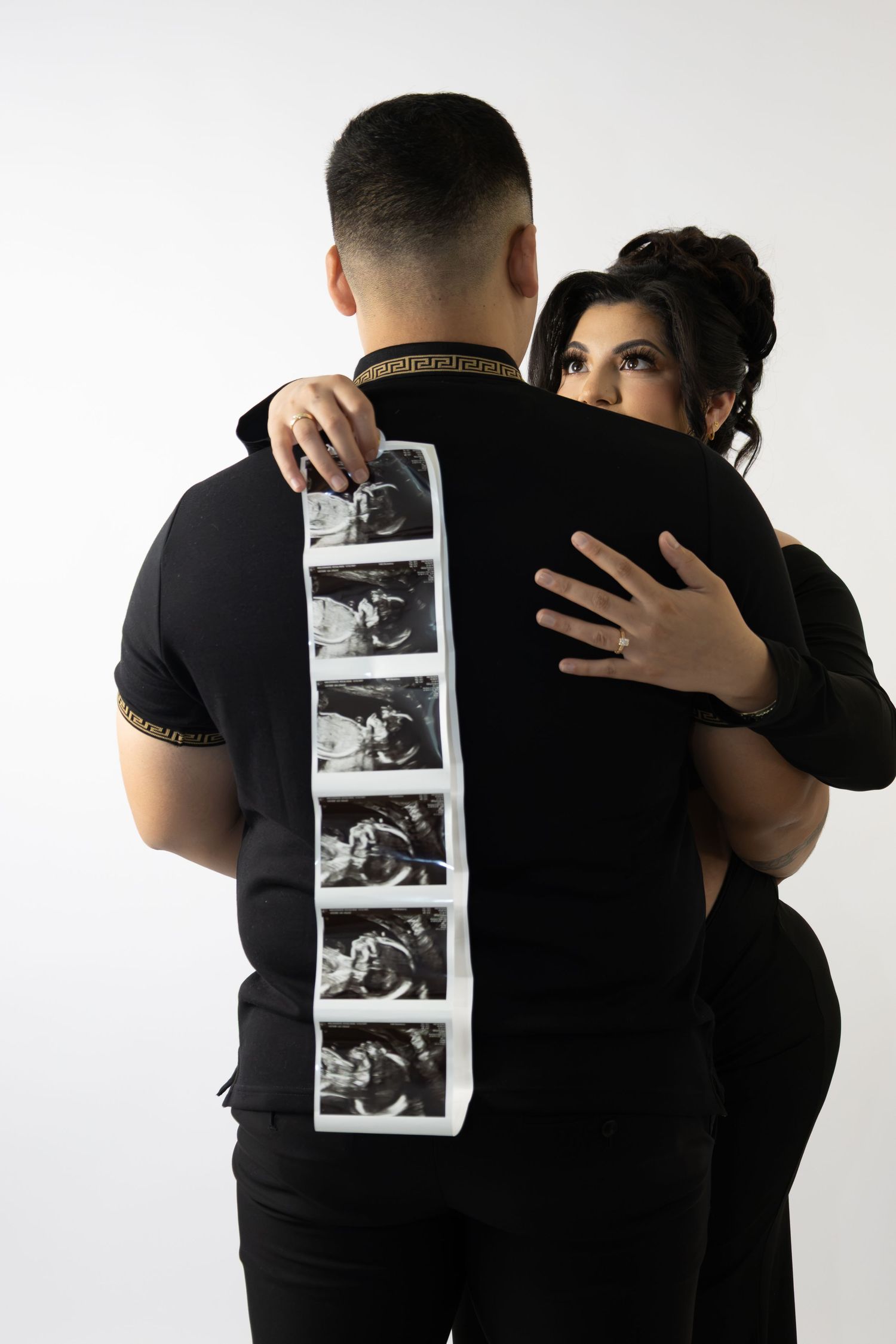 Couple dressed in black embracing while holding ultrasound photo strip against white background.