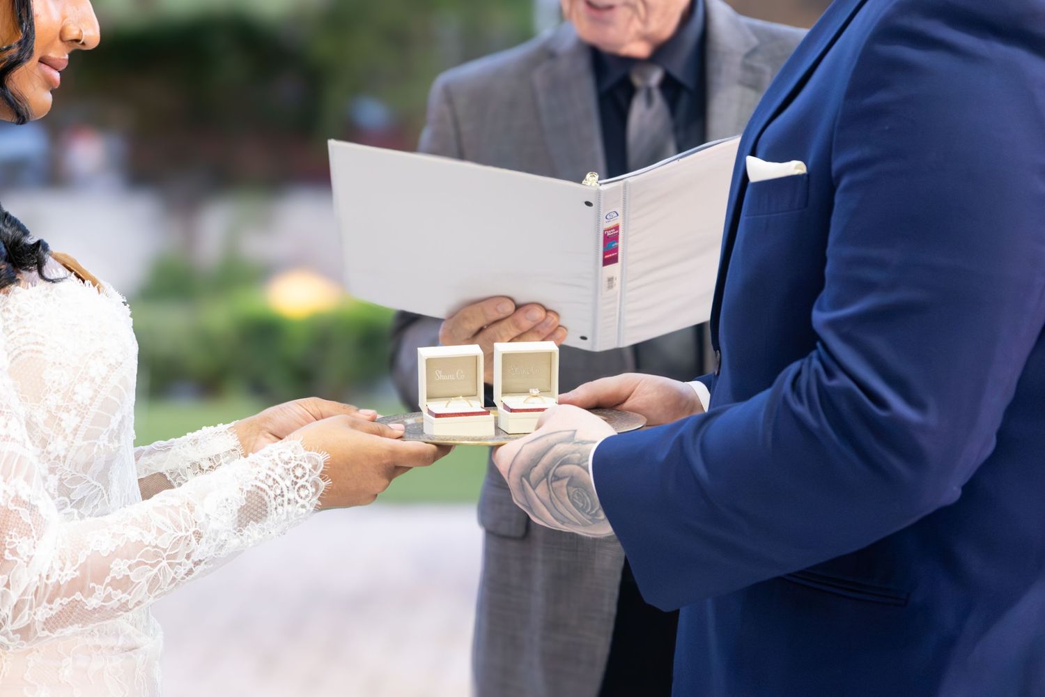 Close-up of wedding ring exchange ceremony with officiant holding book and couple in elegant attire.