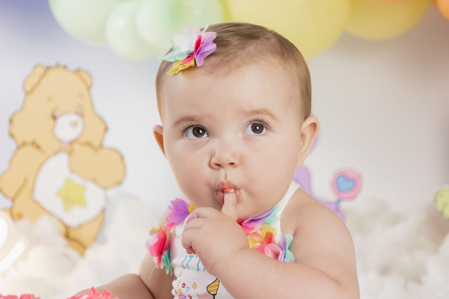 Baby girl in colorful outfit with bow headband at Care Bears themed first birthday cake smash.