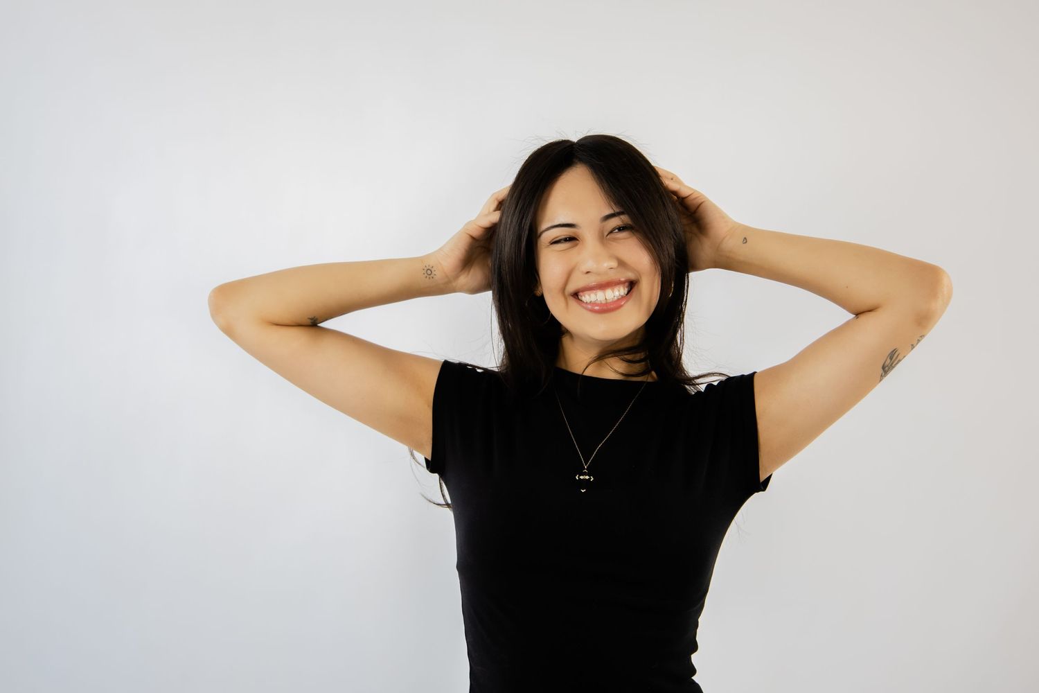 Smiling young woman in black t-shirt with hands in dark hair against white background.