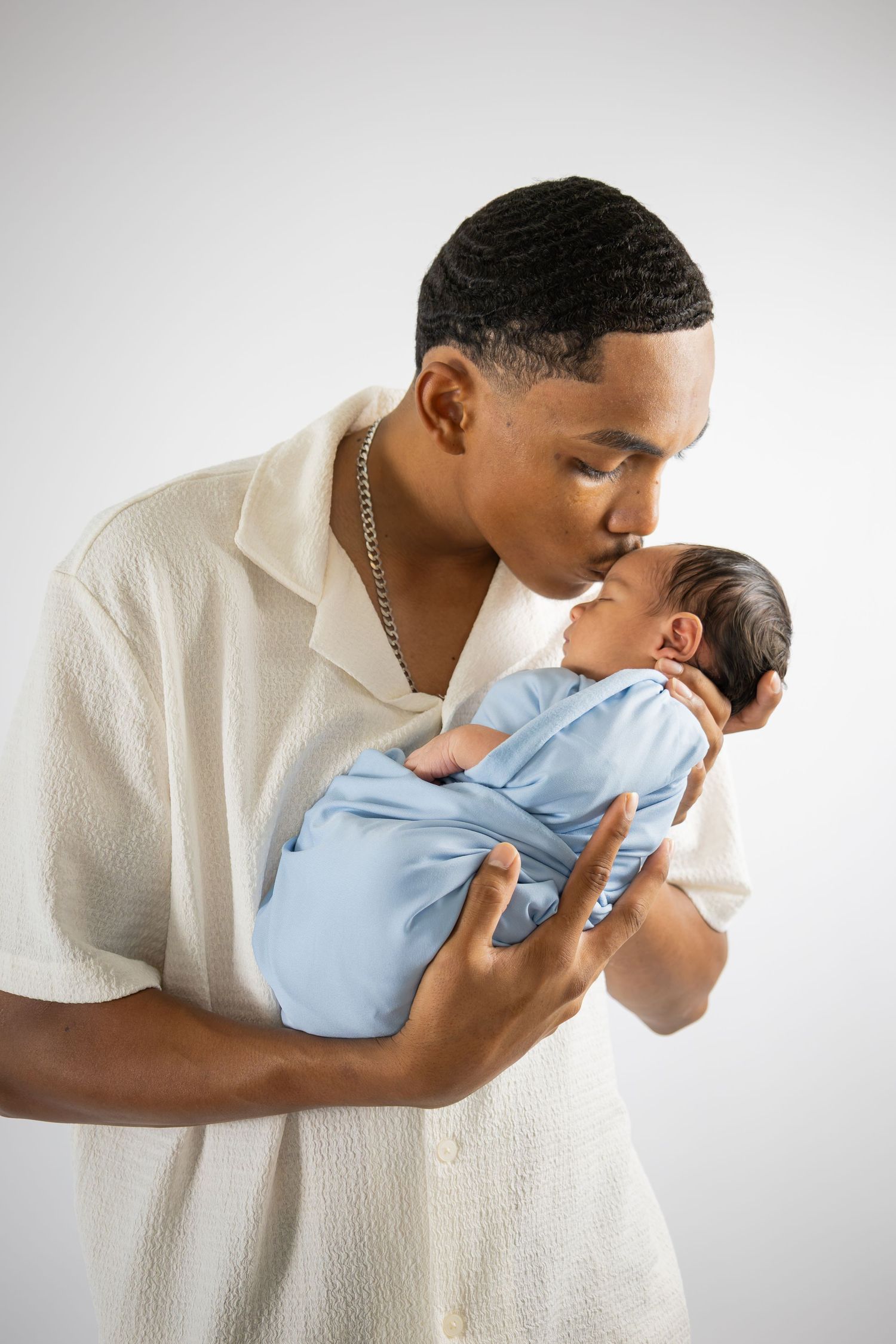 Young African American father kissing his newborn baby wrapped in a blue swaddle blanket.