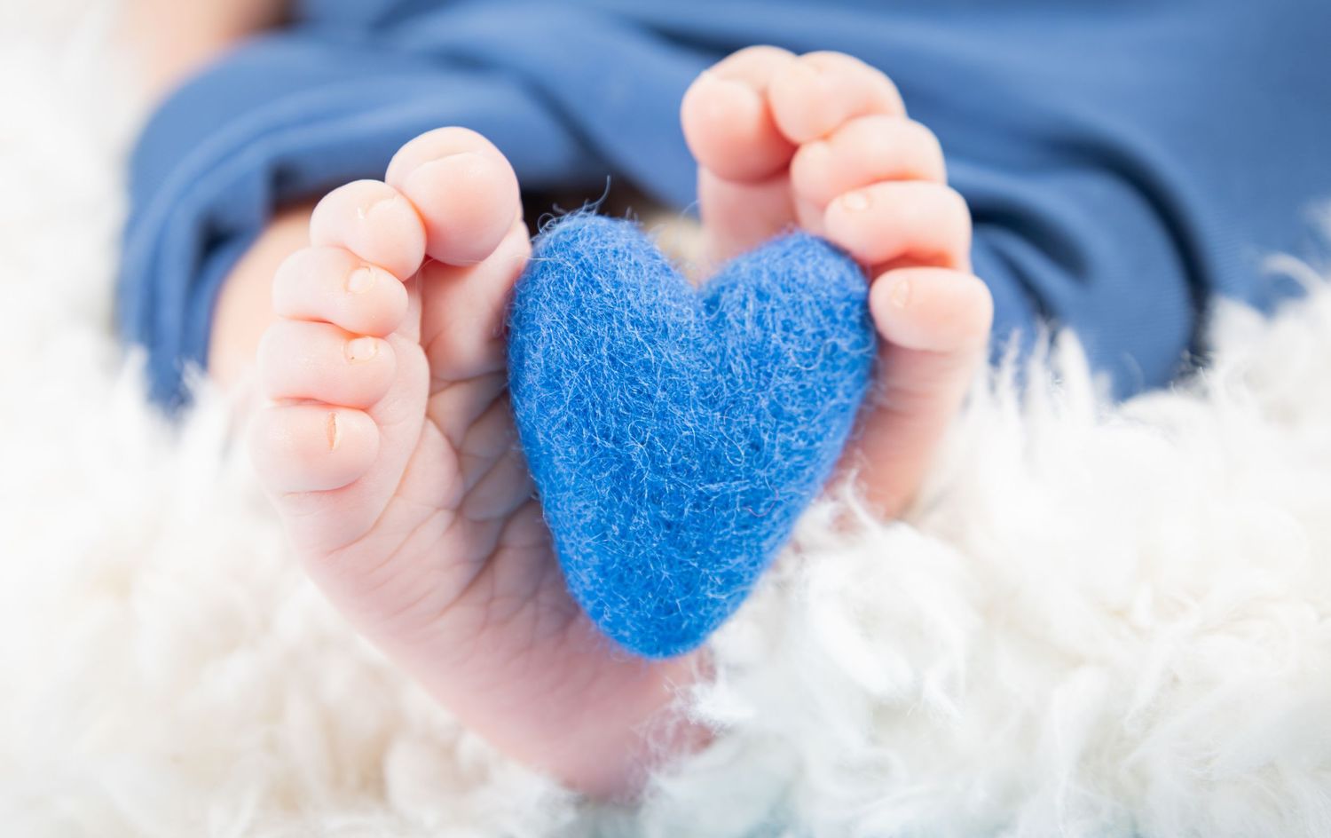 Tiny newborn baby feet holding a small blue felt heart on a soft white fluffy blanket.