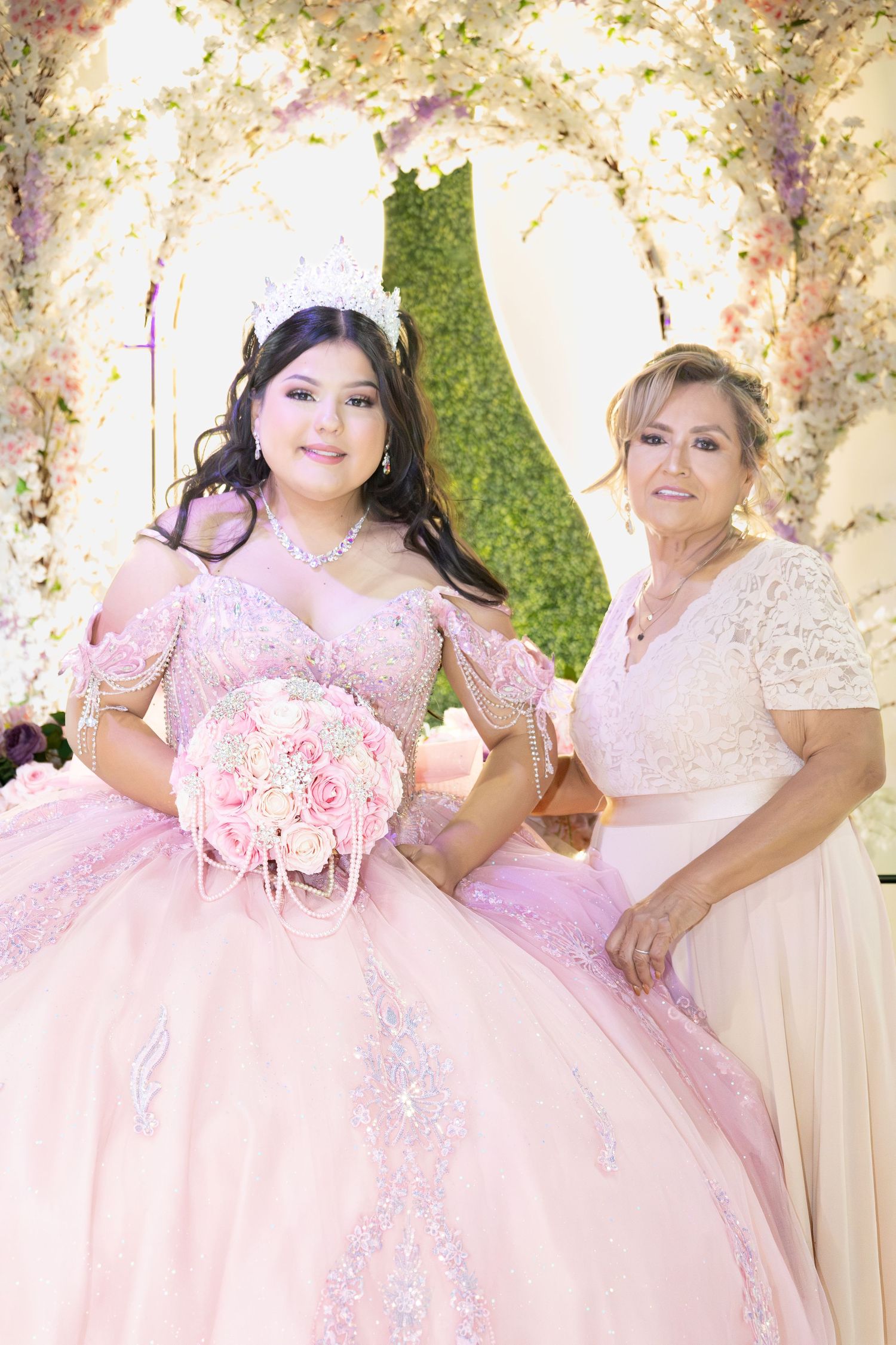 Quinceañera girl in pink ball gown holds rose bouquet beside her mother under floral arch.