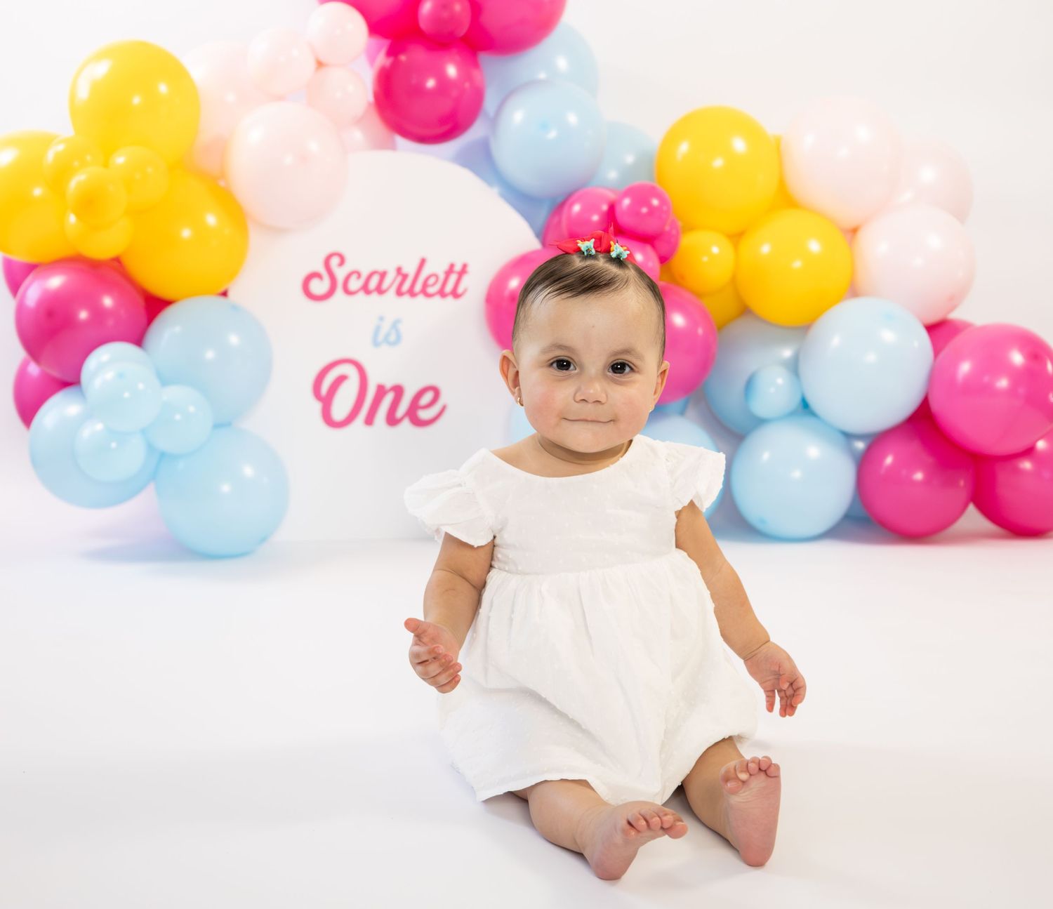 Baby girl in white dress at first birthday celebration with colorful balloon arch backdrop.