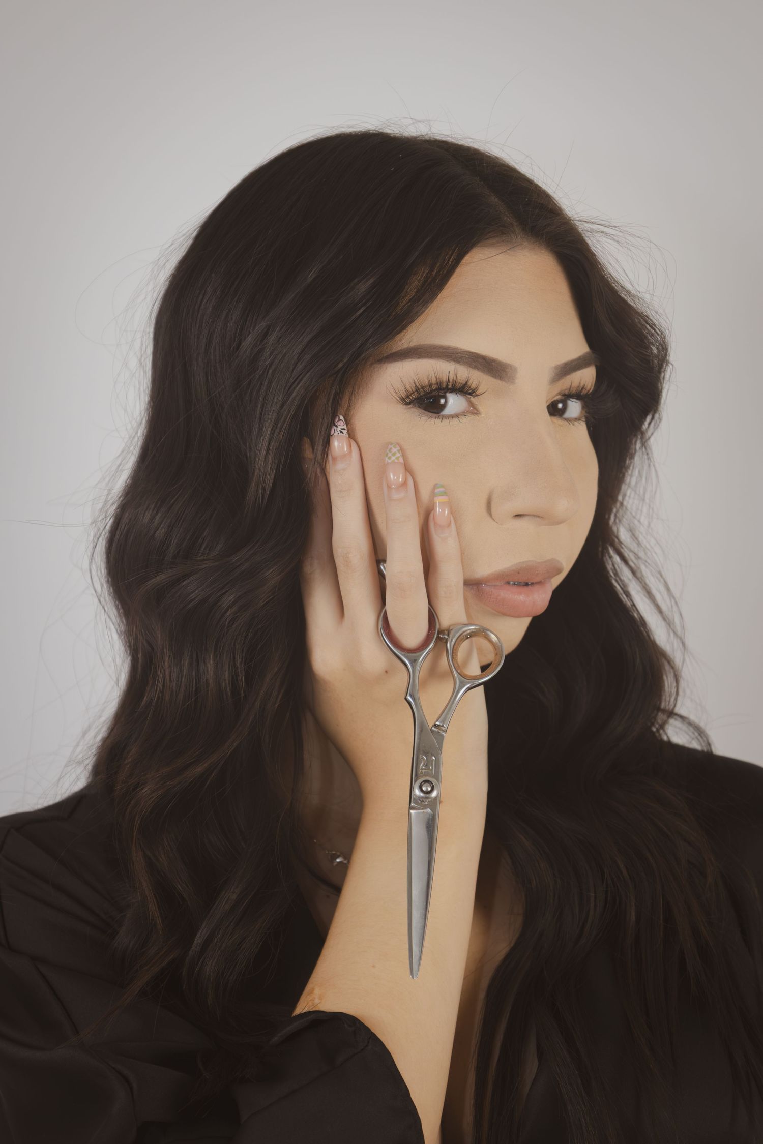 Brunette woman with long wavy hair holding scissors near her face against a light gray background.