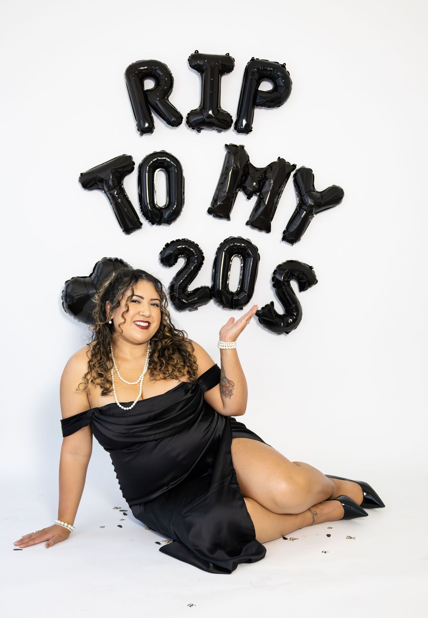 Woman in black dress sitting beside RIP TO MY 20S balloon letters birthday photo.