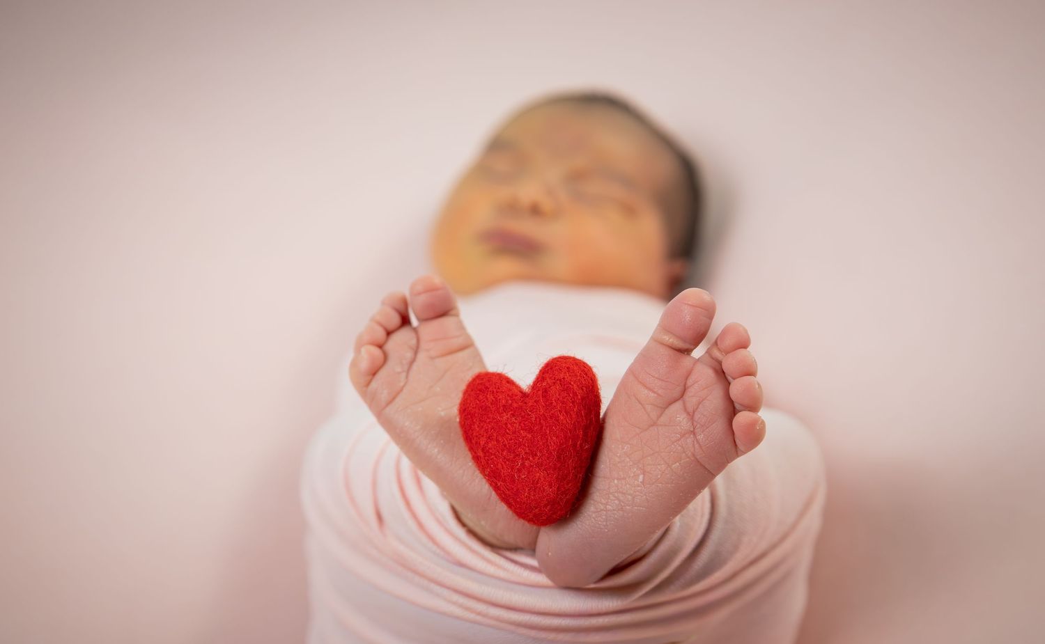 Newborn baby feet holding a small red felt heart while swaddled in a pink wrap blanket.