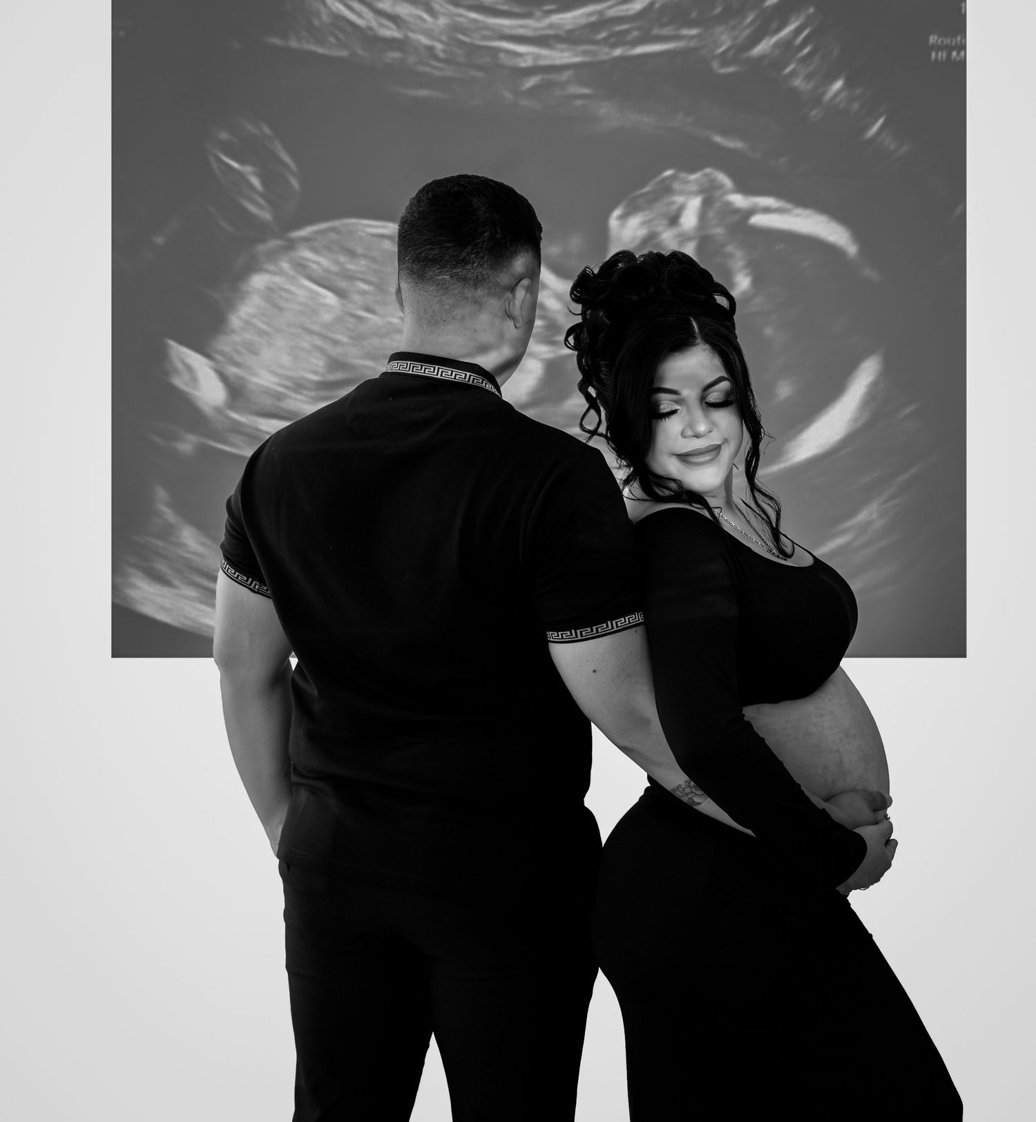 Black and white photo of pregnant couple embracing in front of projected ultrasound image.