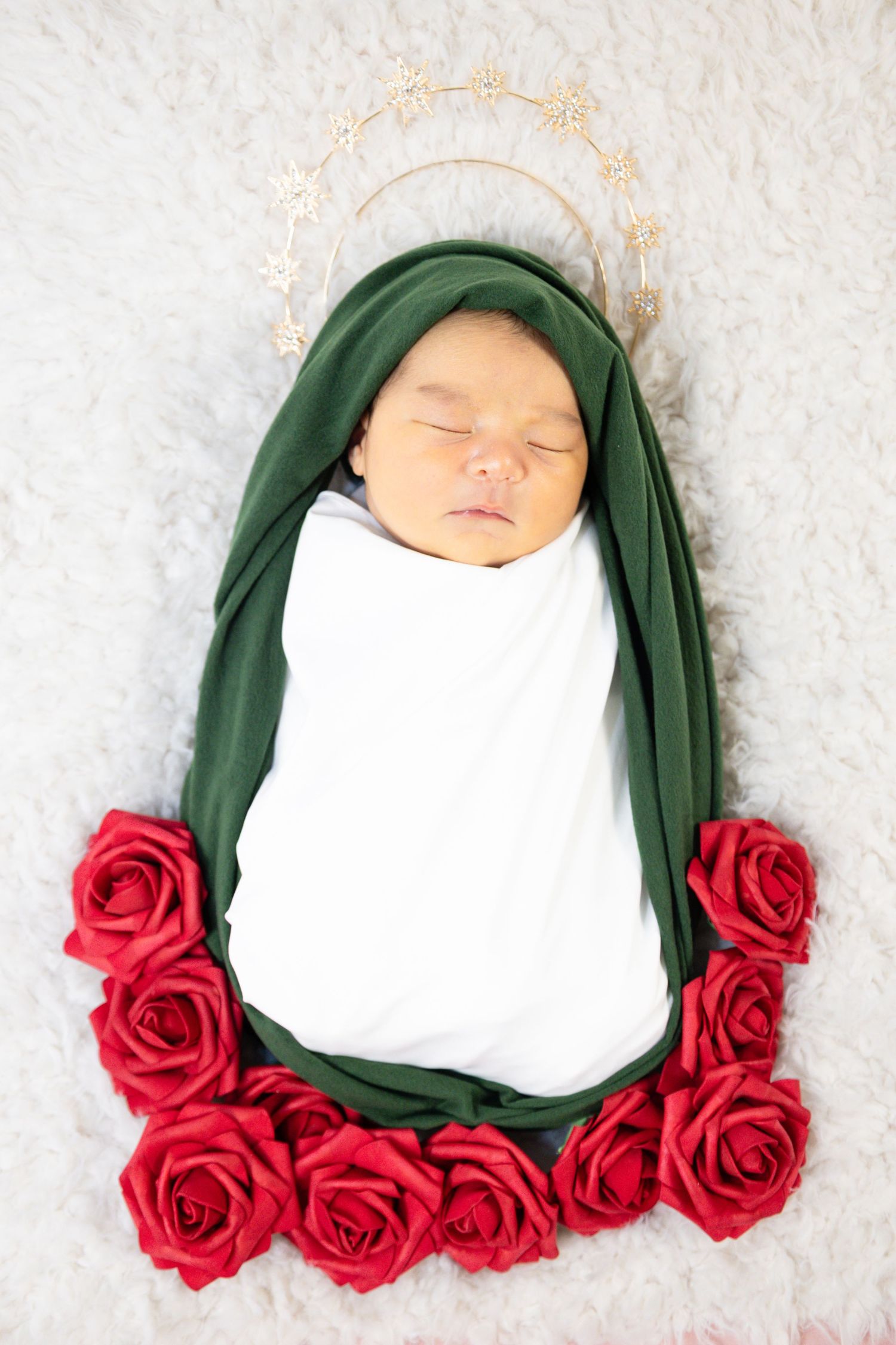 Newborn baby swaddled in white wrap with green cloth and red roses, wearing gold halo, styled as Virgin Mary.