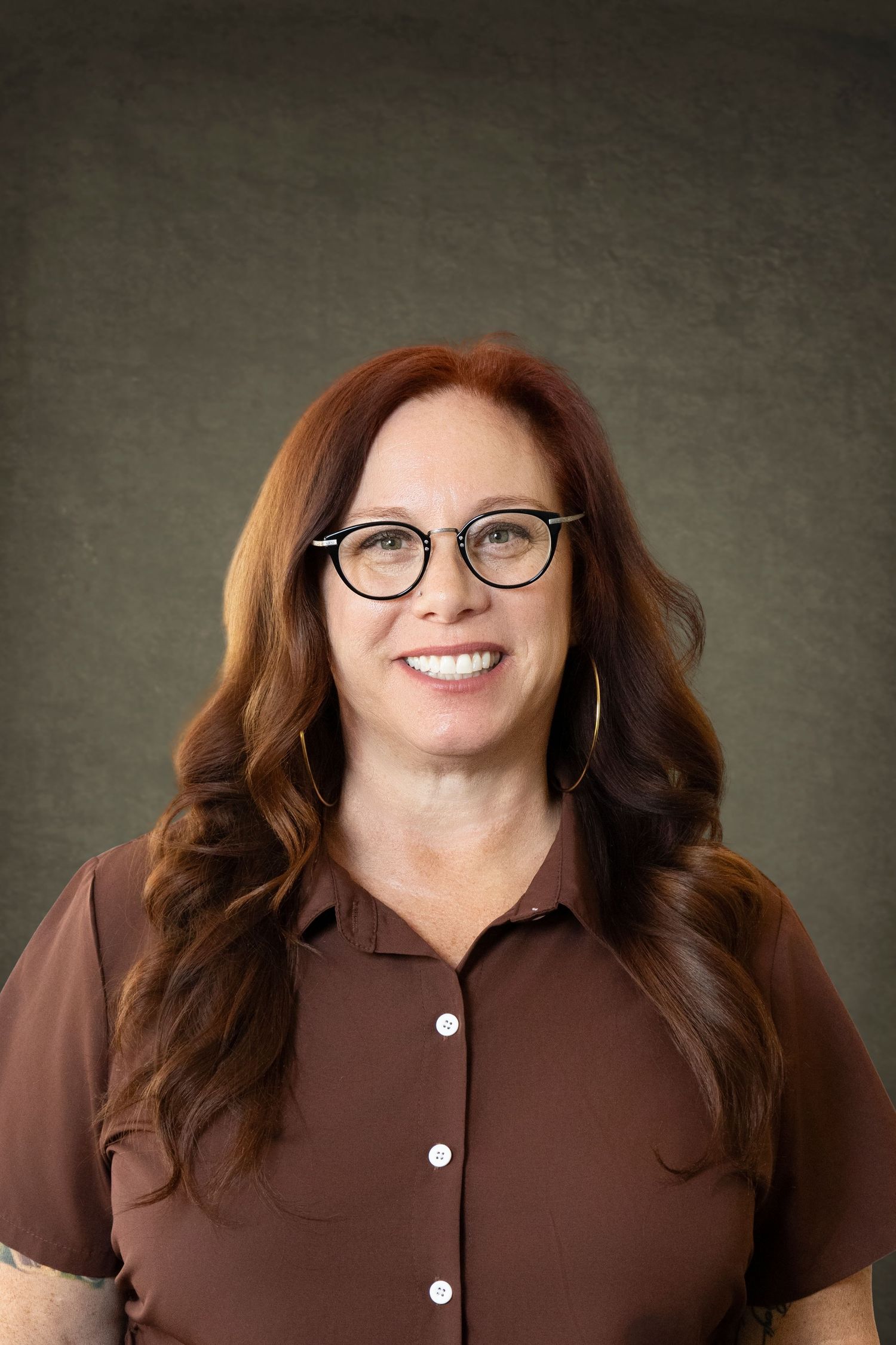 Smiling woman with red hair and glasses wearing a brown shirt poses for a professional headshot photo.