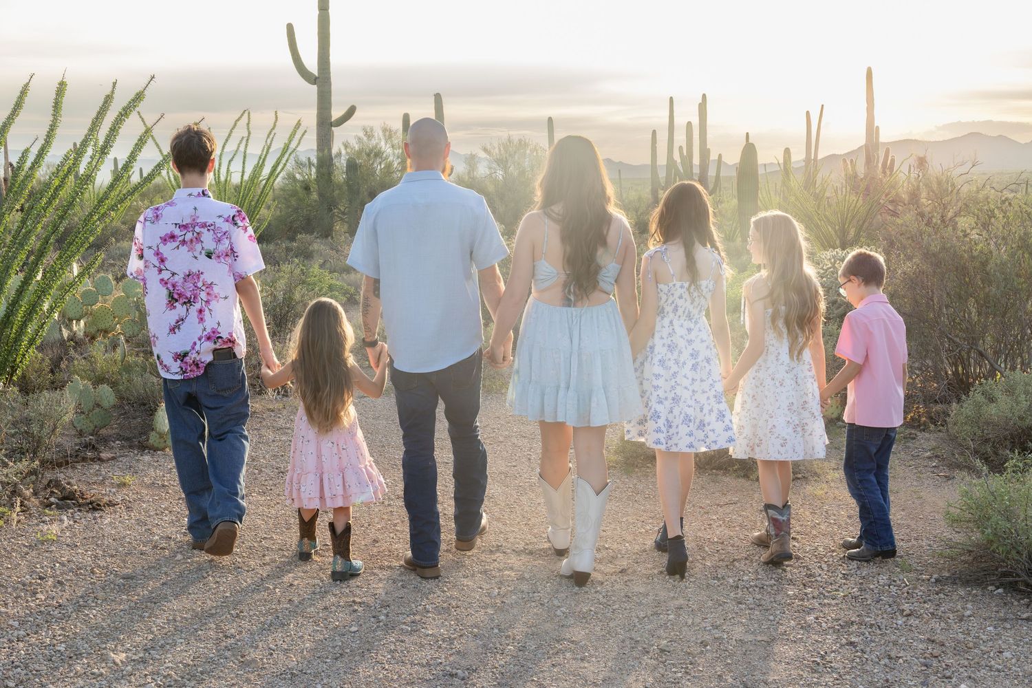 Family of seven walking hand-in-hand along a desert path surrounded by saguaro cacti at sunset.