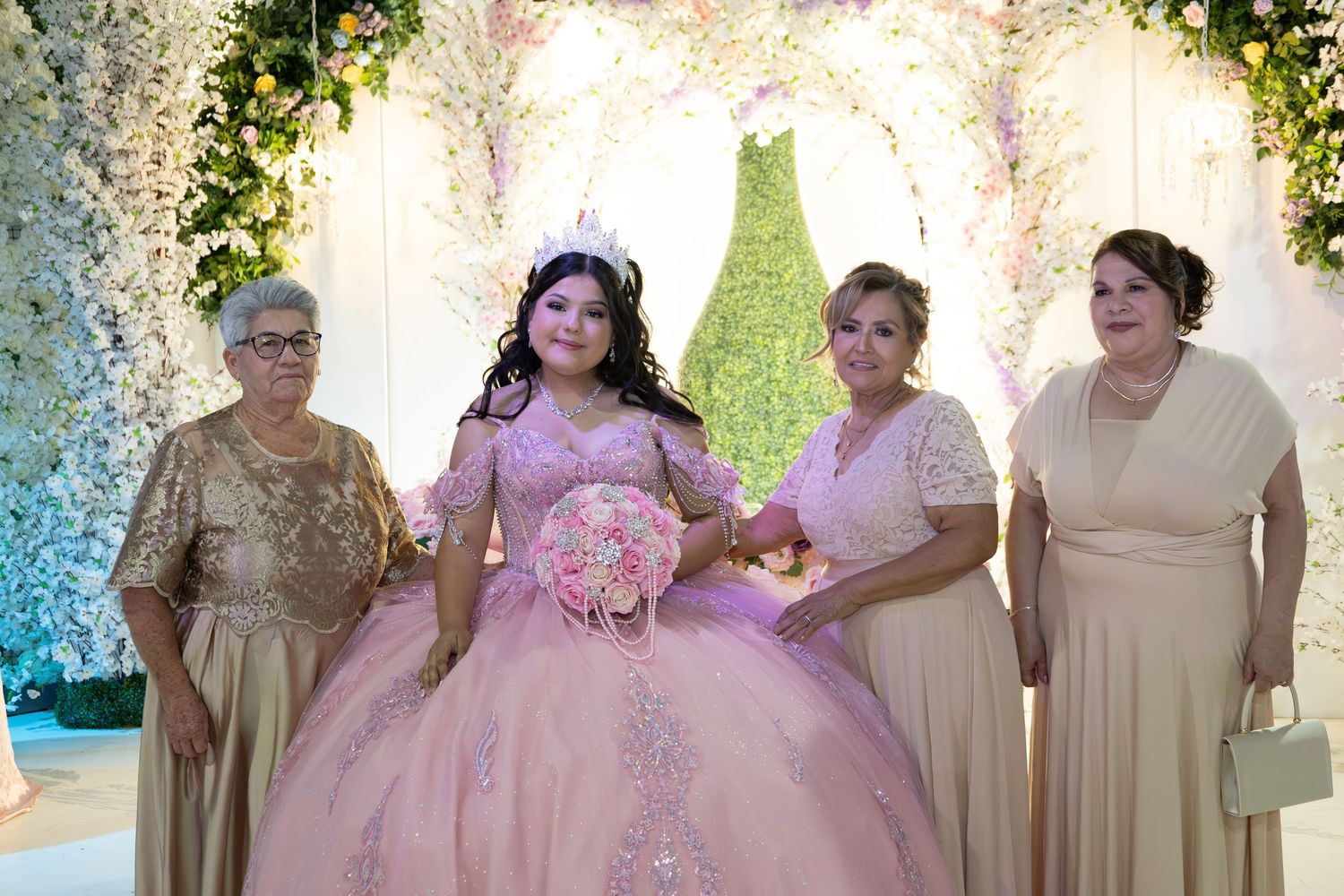 Young girl in a pink quinceañera ball gown poses with three women in front of a floral backdrop.