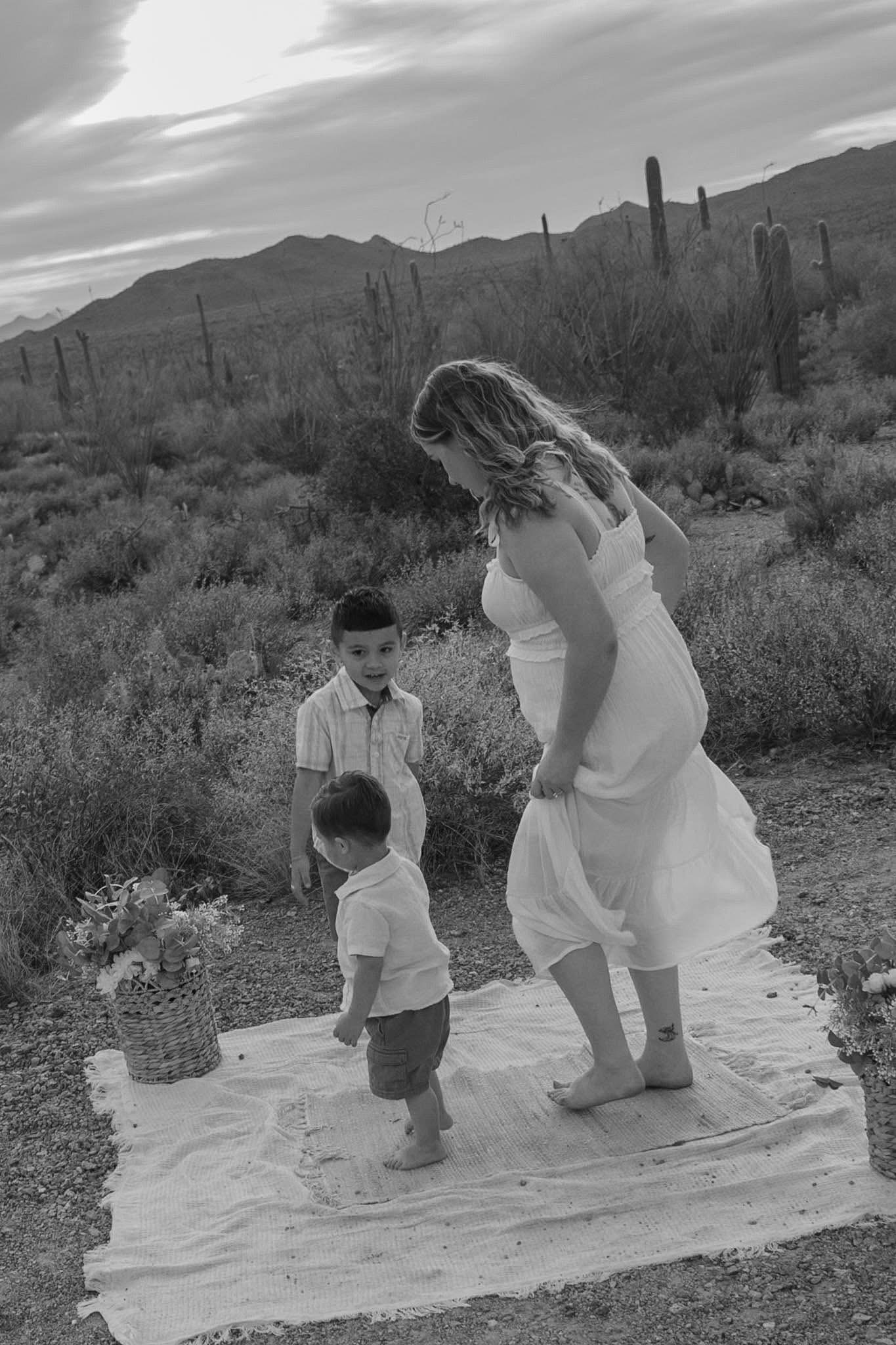 Black and white photo of mother in white dress with two young sons on blanket in desert.