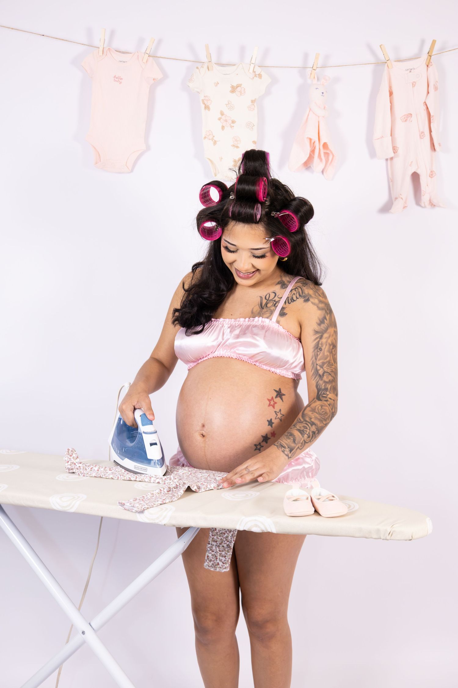 Tattooed pregnant woman in pink lingerie with hair curlers ironing baby clothes with laundry hanging in background.