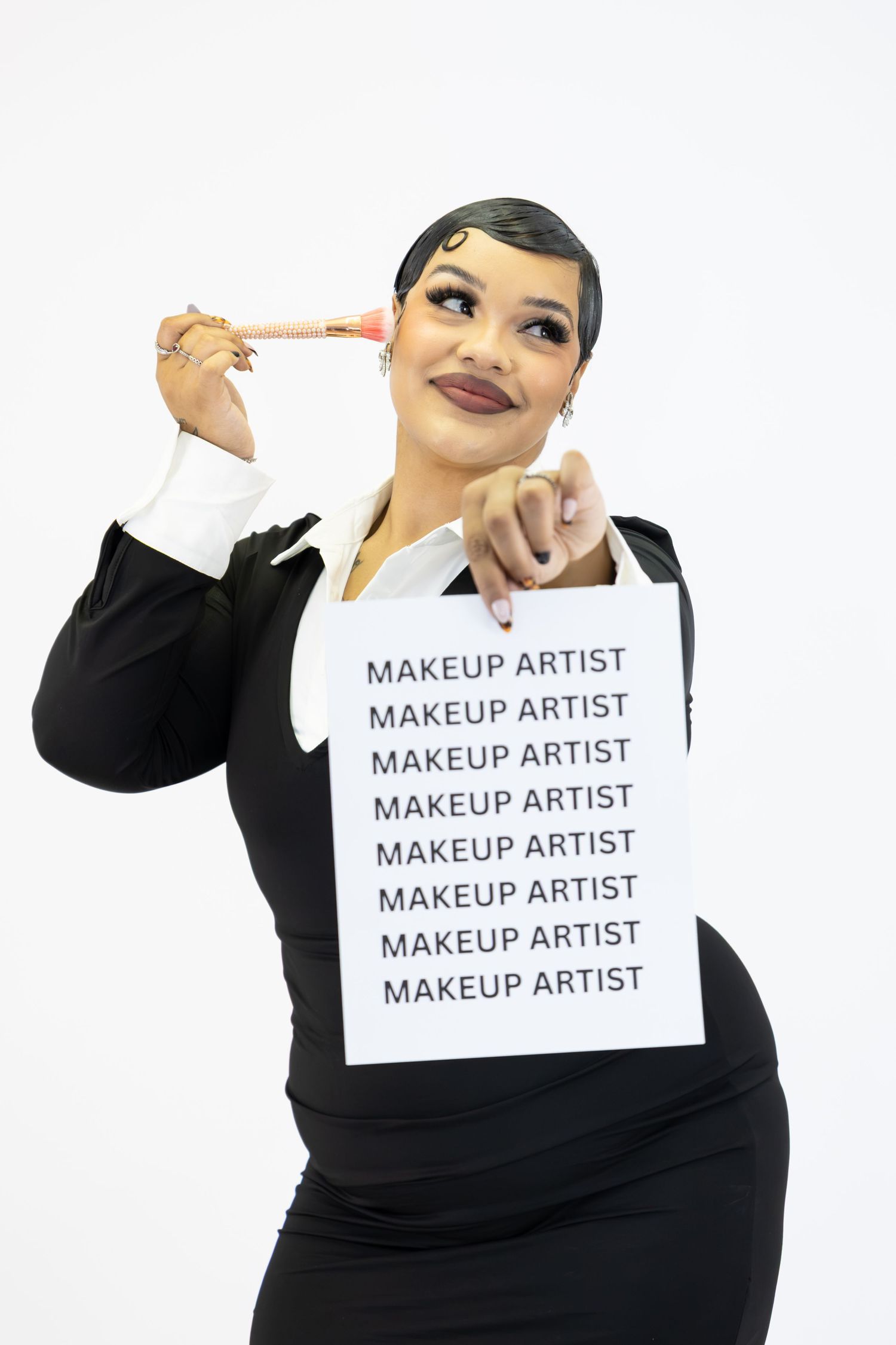A makeup artist in a black suit holds a sign reading MAKEUP ARTIST repeatedly while pointing a brush at her temple.