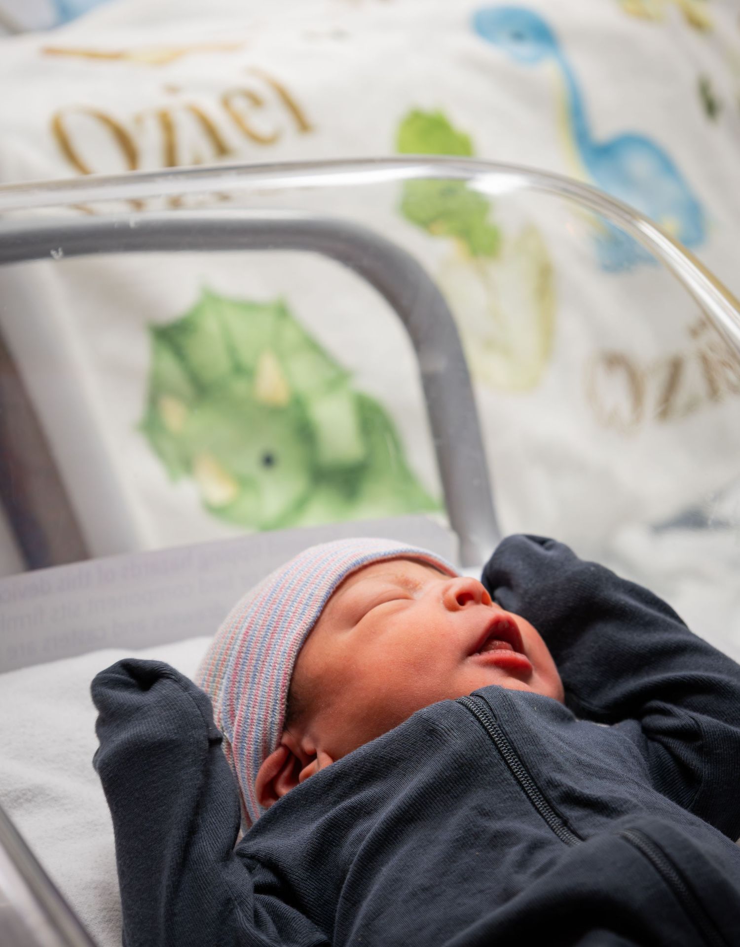 Newborn baby in a hospital bassinet wearing a striped hat beside a dinosaur-themed blanket.