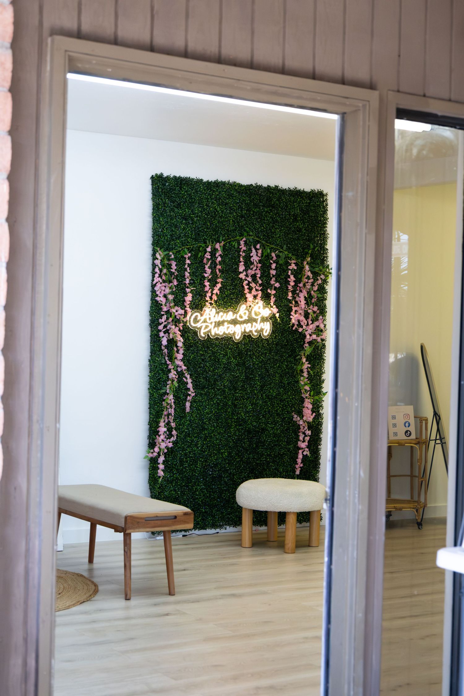 Photography studio interior with green boxwood wall, neon sign, pink florals, and minimalist white furniture.