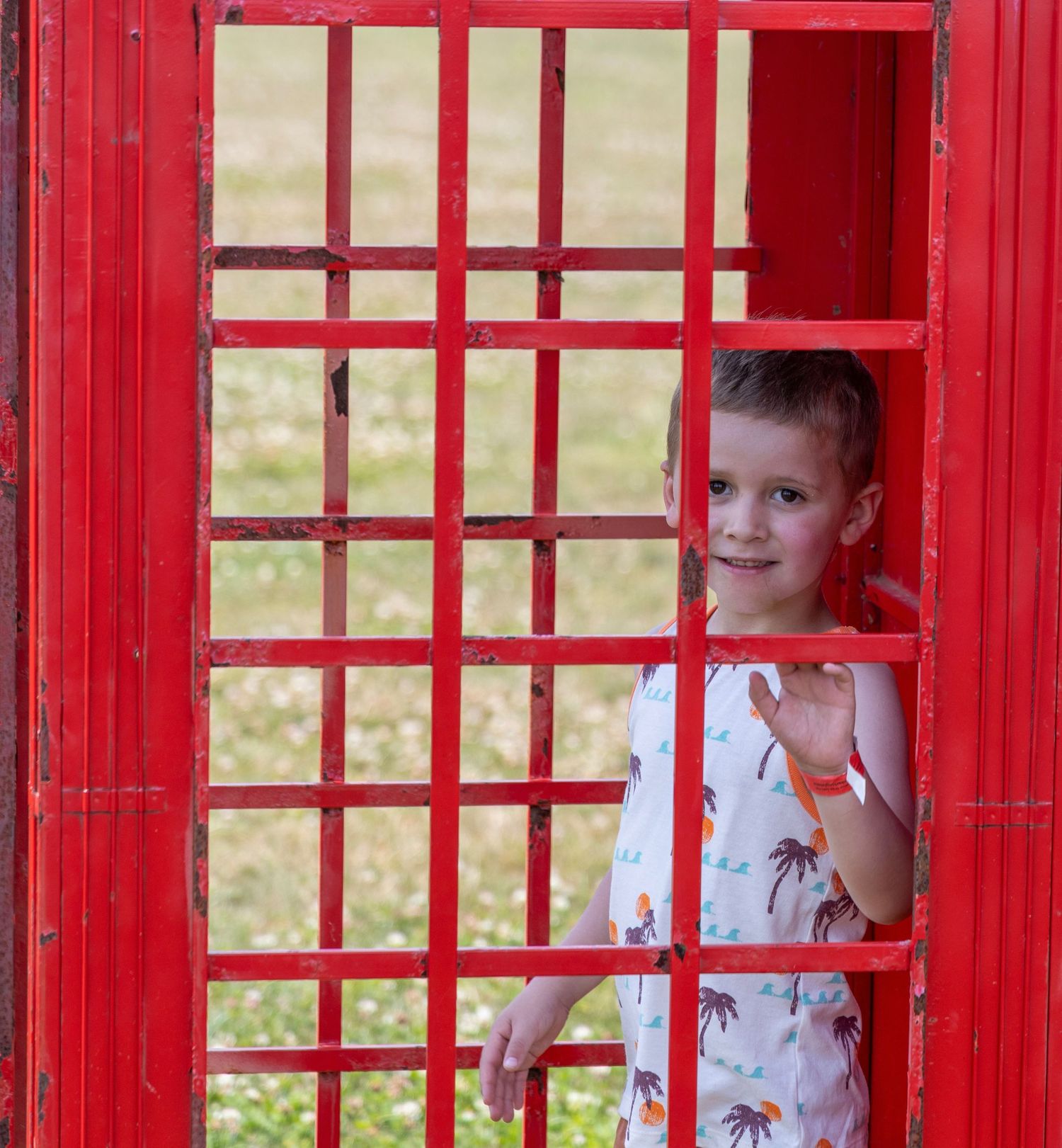 Smiling young boy waving from inside a classic red British telephone box outdoors.