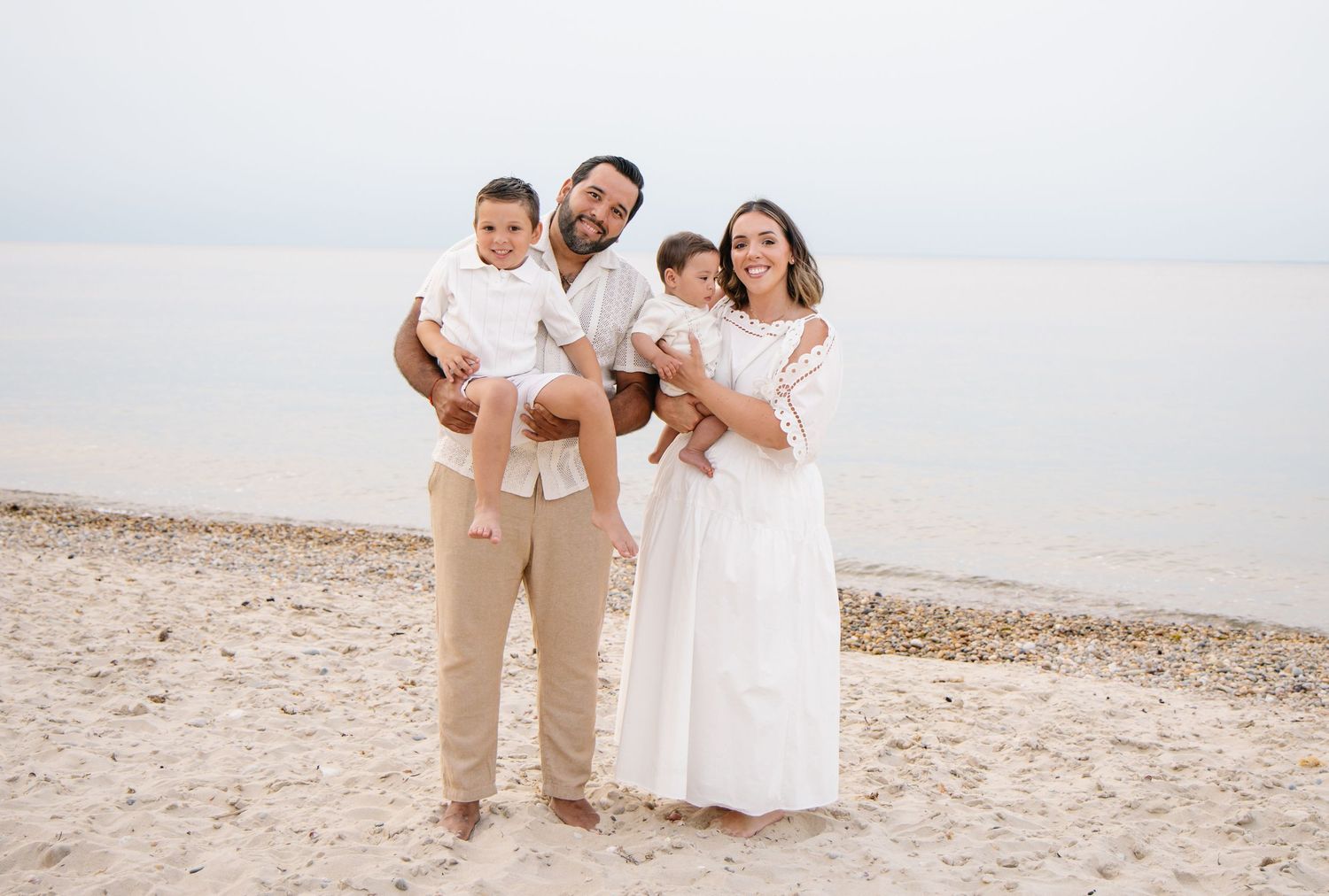 Smiling parents each holding a young child on a serene sandy beach.