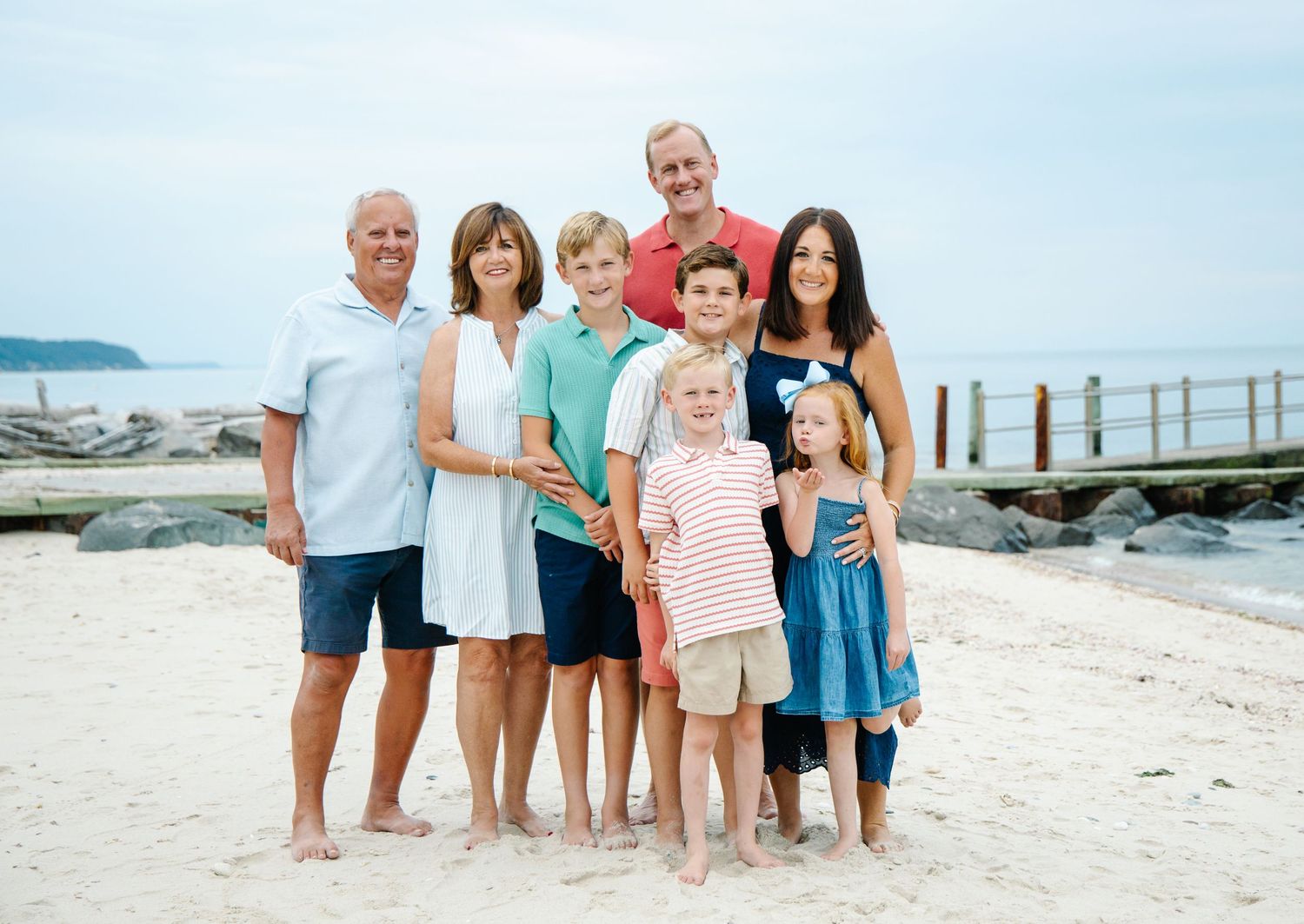 Extended family of eight posing barefoot on sandy beach with ocean backdrop.
