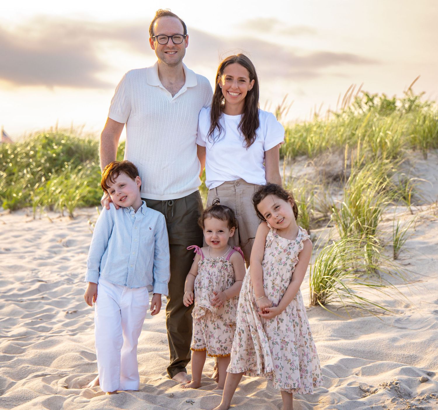 Happy family of five posing together on a sandy beach at sunset, with two adults and three young children smiling. Hamptons beach session.