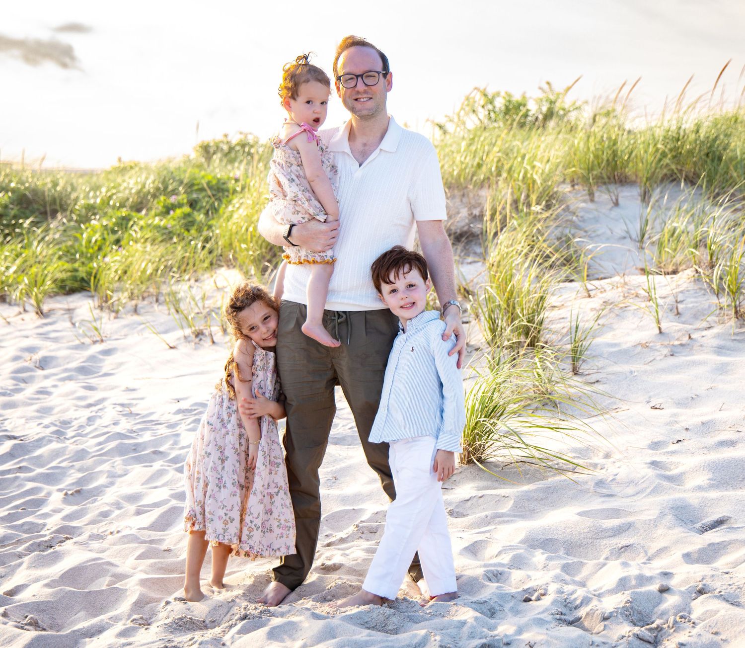 A father poses with three young children on a sandy beach with dunes and green grass in the background. family photographer. Hamptons photo