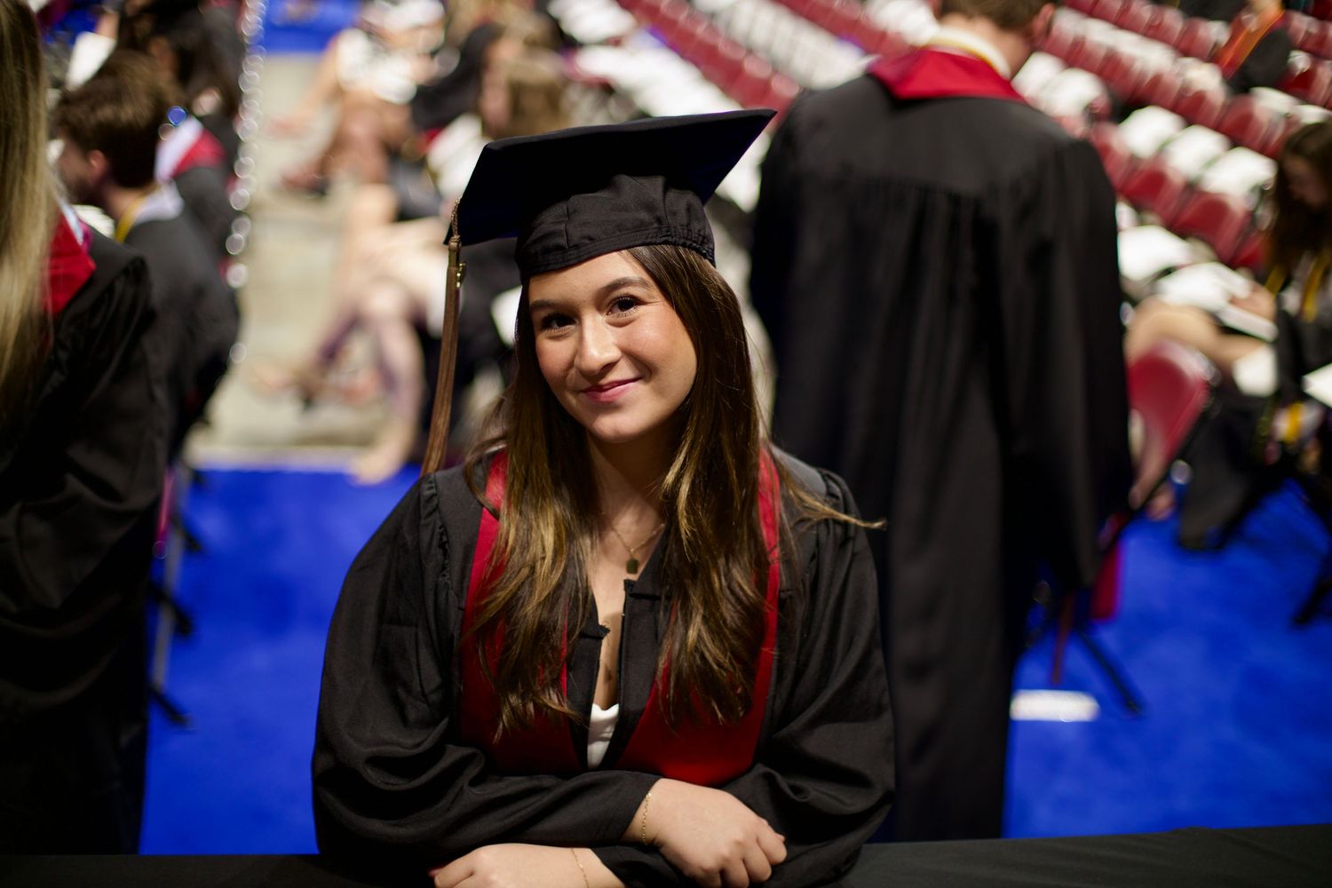 Smiling female graduate in black cap and gown with red trim at graduation ceremony.