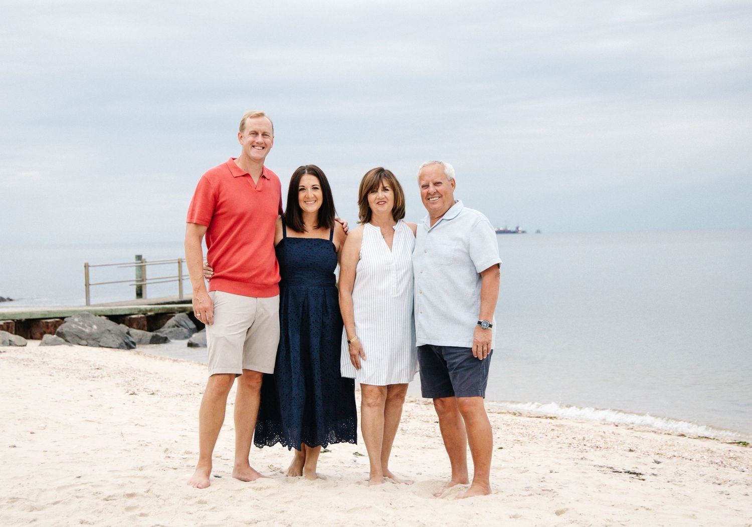 Two couples smiling together on sandy beach with calm ocean water behind them.