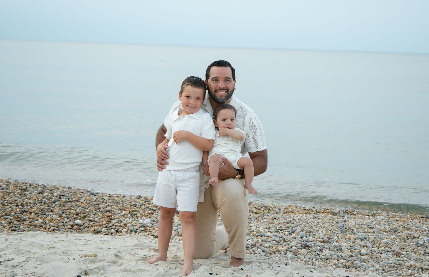 Father in white shirt poses with two young sons on a rocky beach shore with calm water behind.