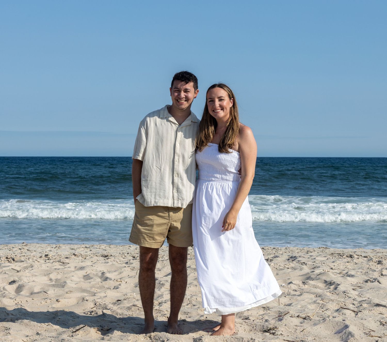 Young couple smiling together on sandy beach, man in white shirt and khaki shorts, woman in white dress.