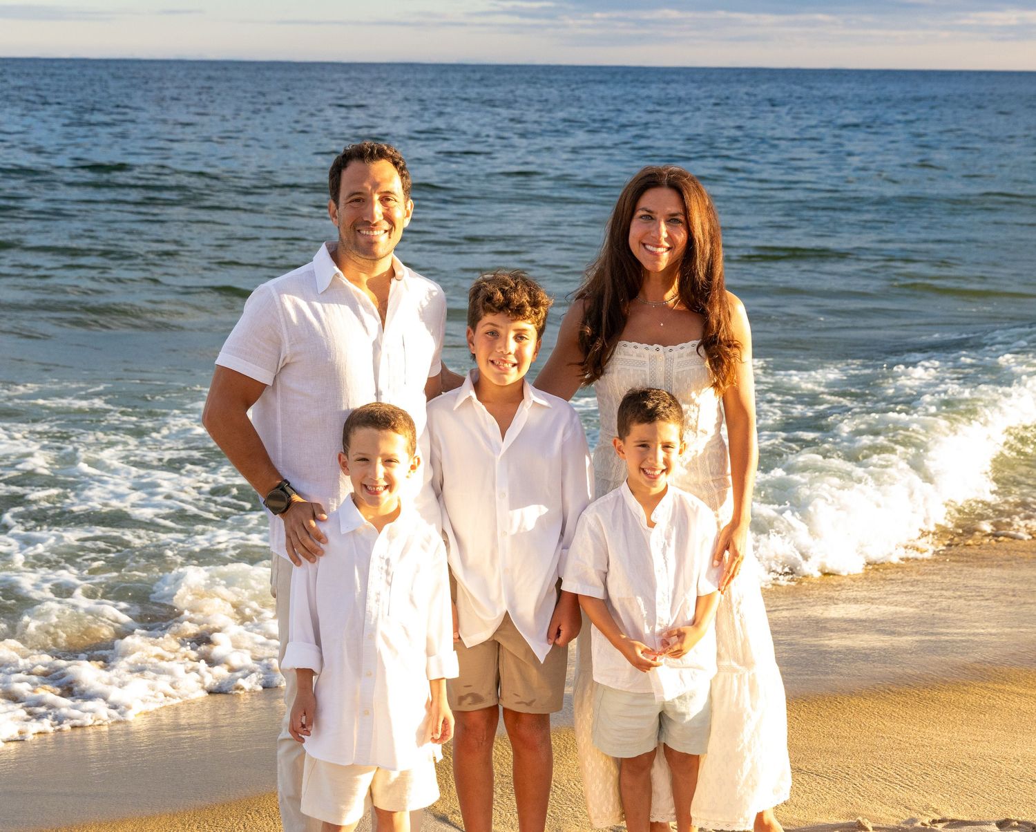 Family of five posing on a sunny beach in white outfits with ocean waves in the background. Family Photographer. East Hampton Beach.