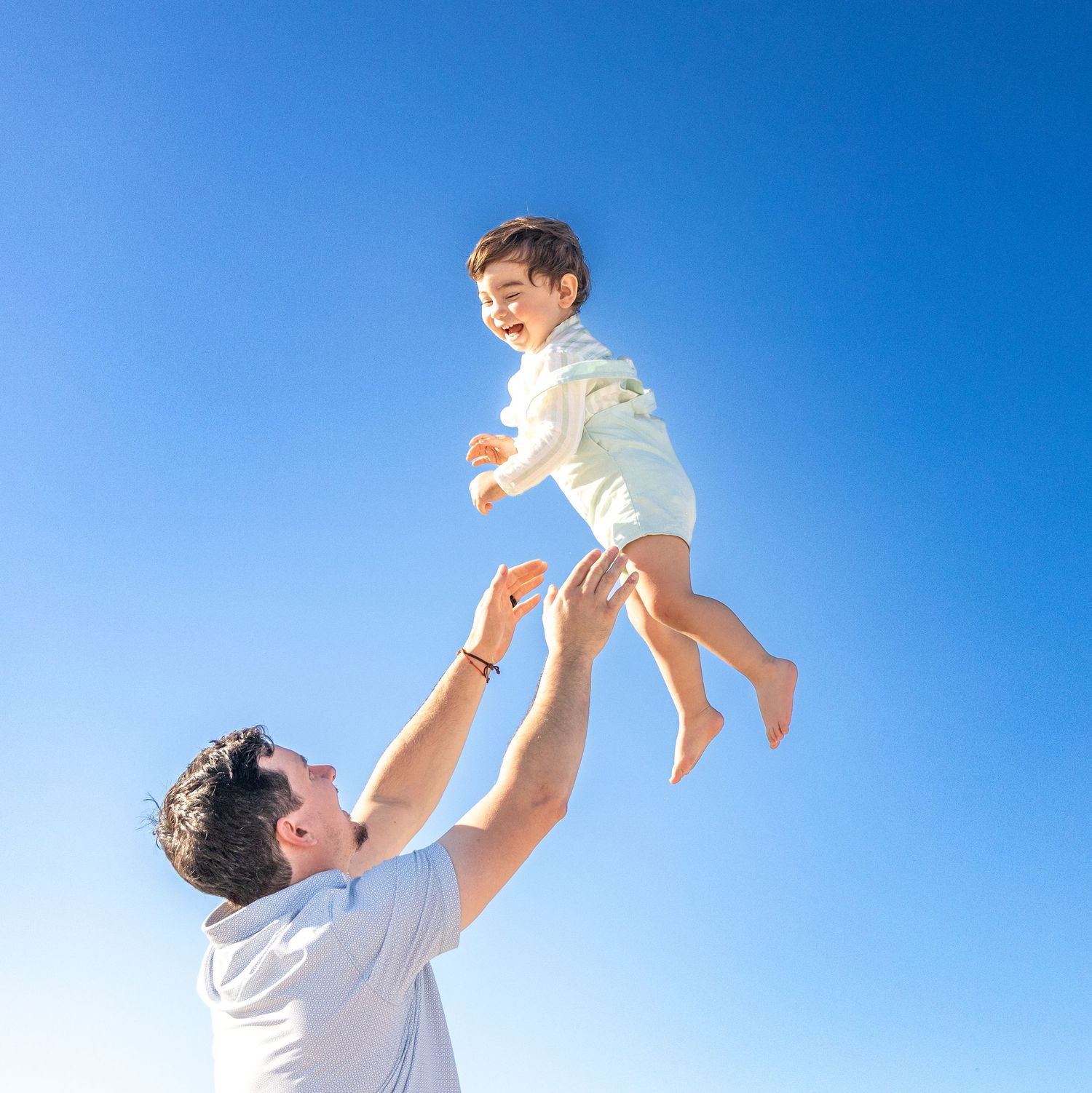 A joyful father tosses his laughing toddler high into a clear blue sky on a sunny day. Hamptons photographer.family photographer Long Island