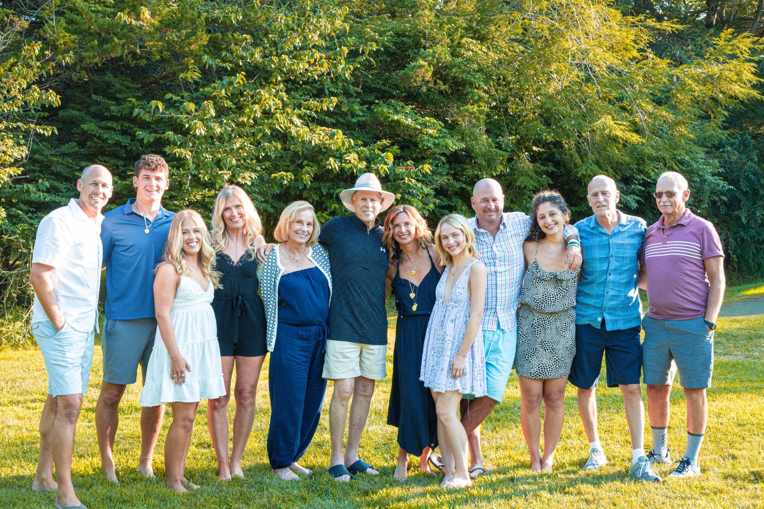 Large family group of eleven adults posing together on green lawn with trees behind them.
