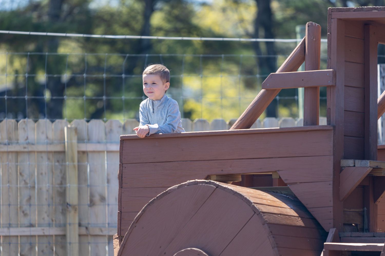 Young toddler boy standing on wooden playground structure outdoors on sunny day.