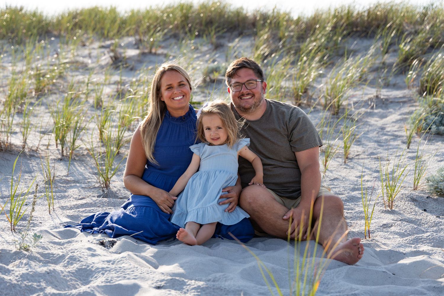 Smiling family of three sitting in beach sand dunes with sea grass, parents flanking young daughter.