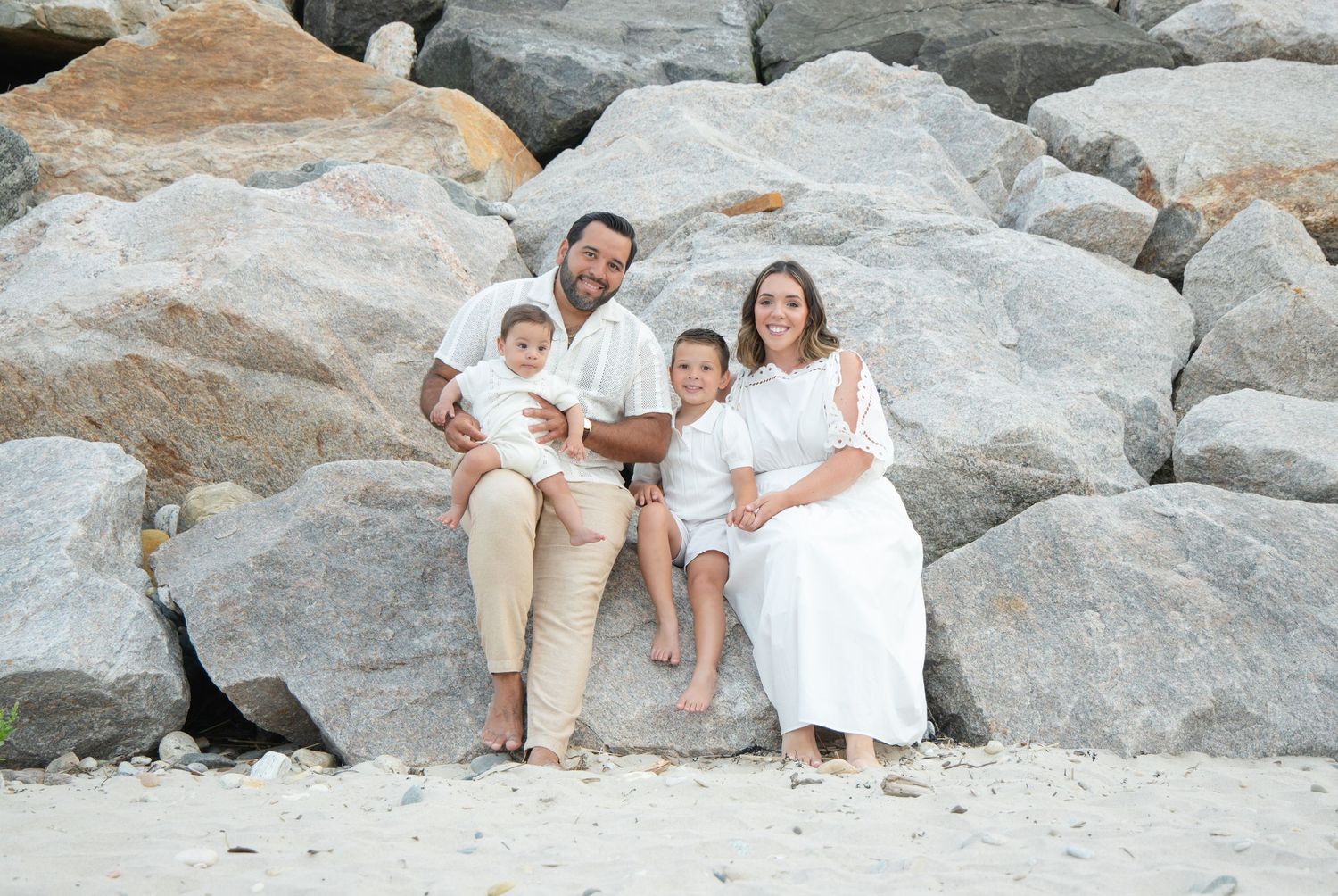 Smiling family of four in white outfits sitting together on large rocks at the beach.