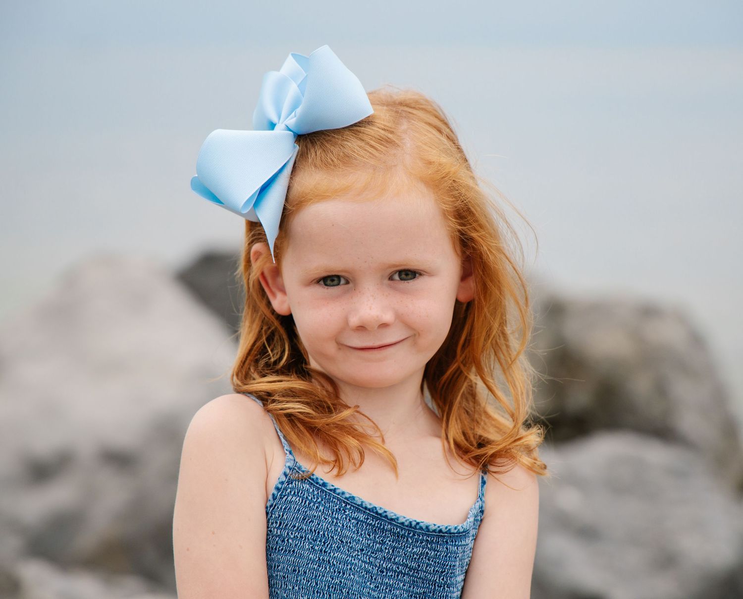 Adorable red-haired girl with light blue bow smiling at beach portrait session.