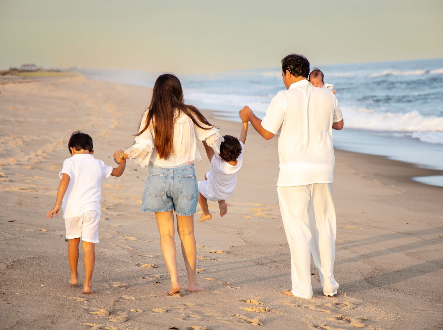 Family of five walking barefoot on a sandy beach at sunset, wearing white outfits, holding hands near ocean waves. Hamptons family photo