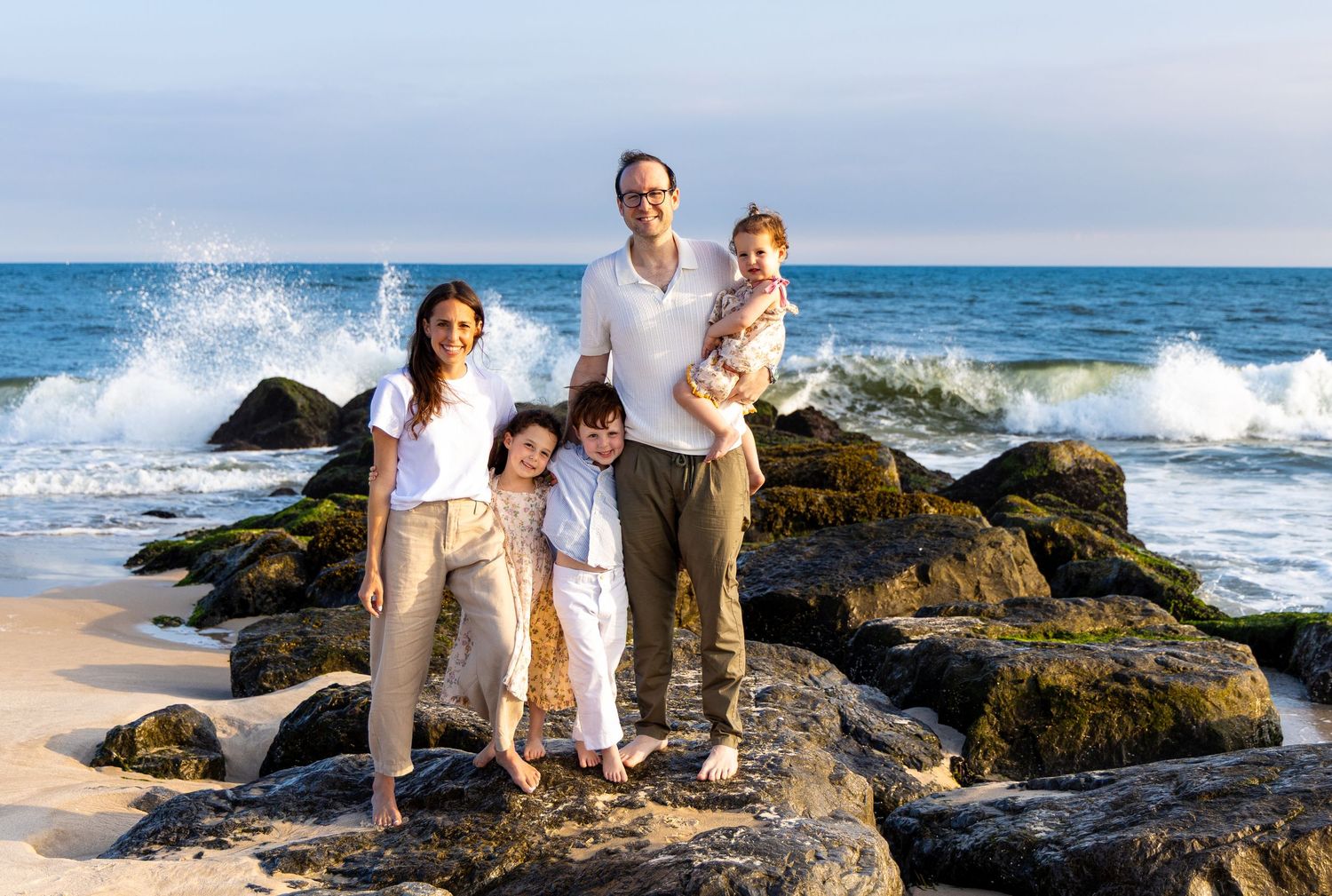 Family of four posing on rocky beach with ocean waves crashing behind them on a sunny day. Hamptons photographer. family photographer