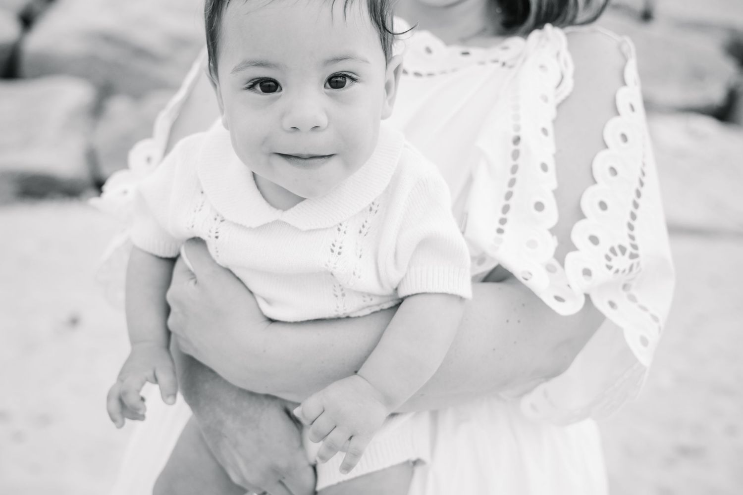 Black and white portrait of a smiling baby in white outfit being held by a woman on beach.